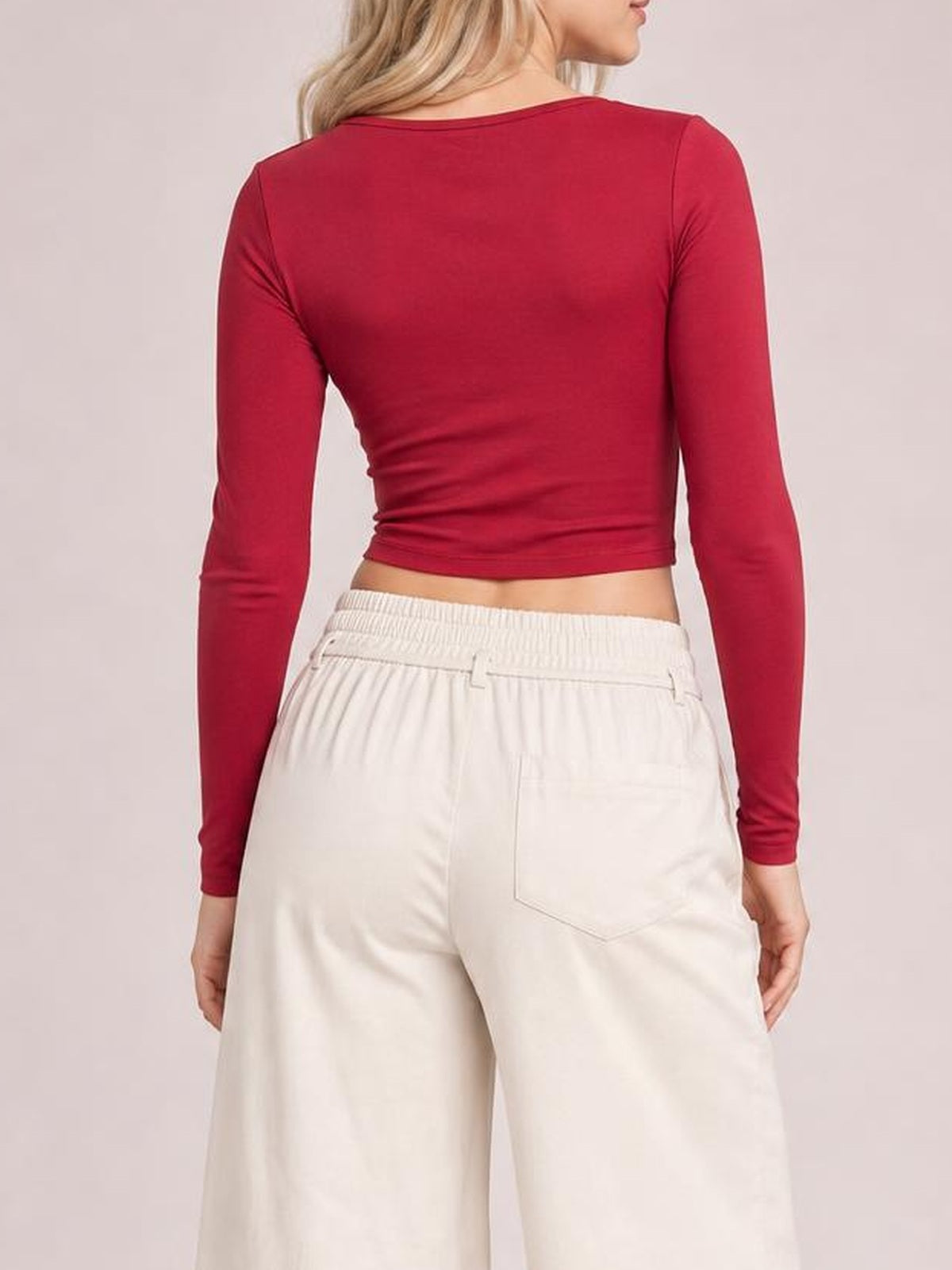 A model stands turned away to show cream high-waisted wide-leg pants with a buttoned waistband, relaxed silhouette, and visible back pockets.