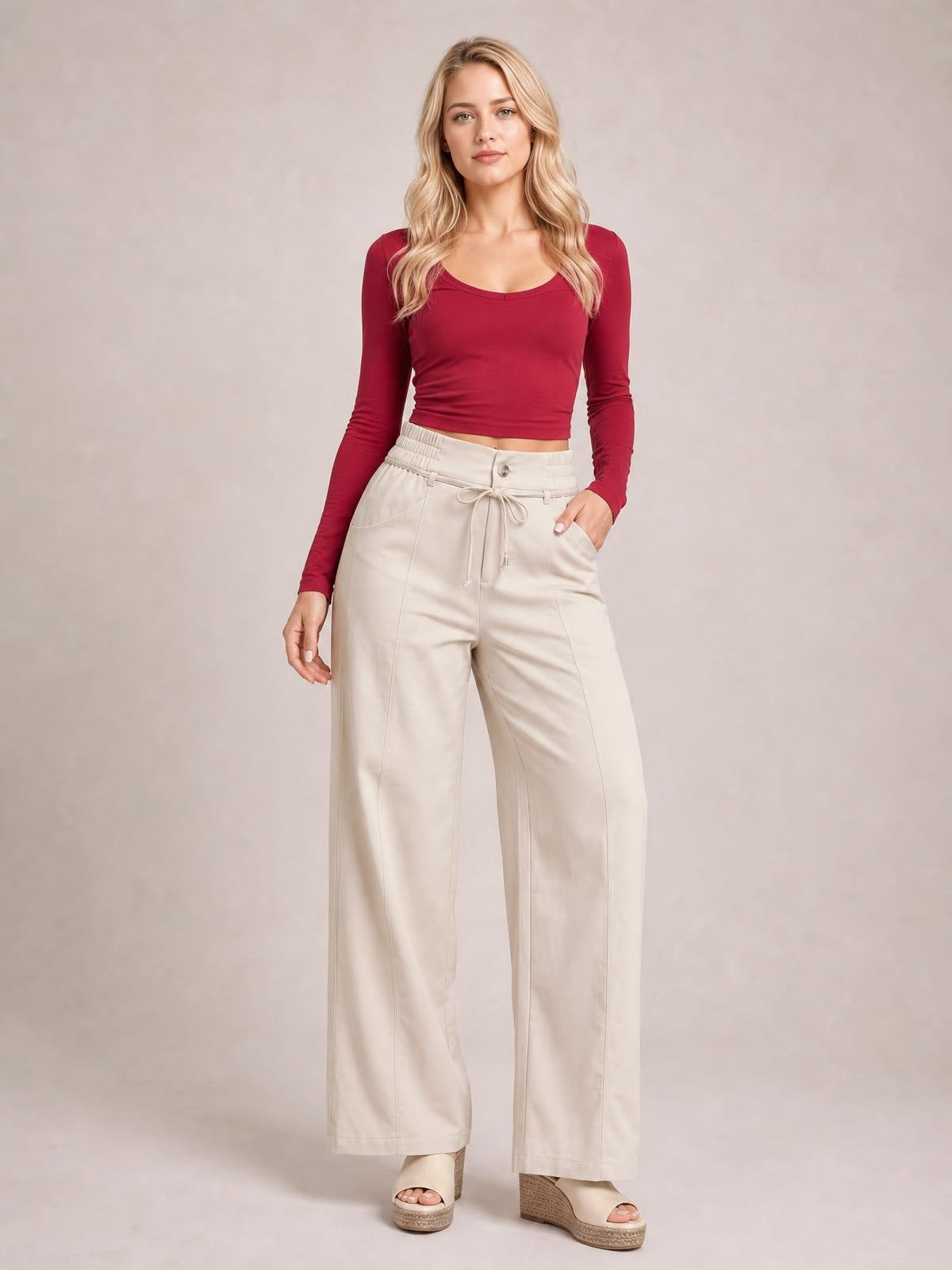 Model stands with one hand in a pocket, wearing cream high-waisted wide-leg pants with a button closure, tied drawstring waist, and long straight seams.