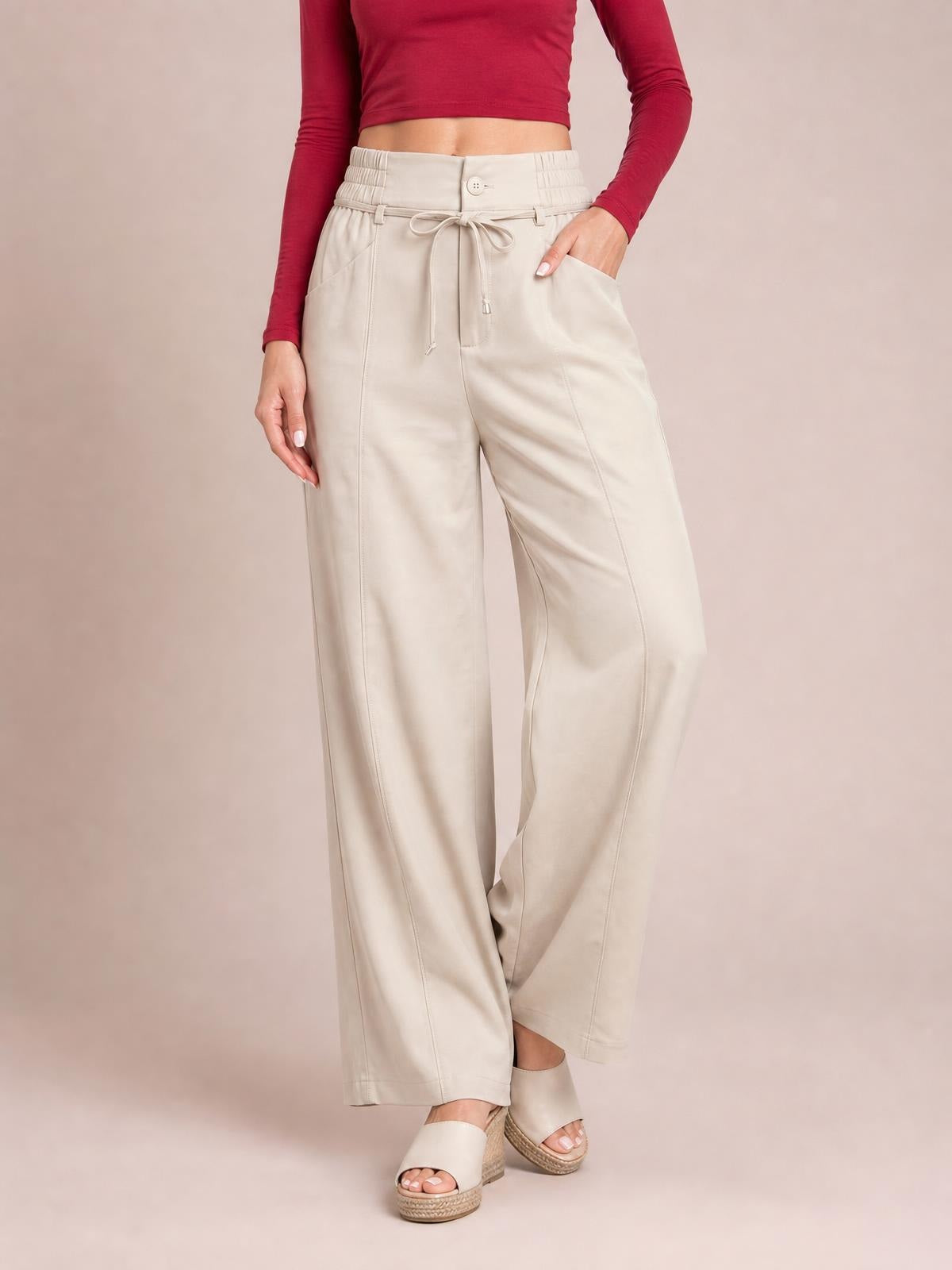 Model wears high-waisted cream wide-leg pants with a button closure, tied drawstring waist, side pockets, and stitched panel construction.