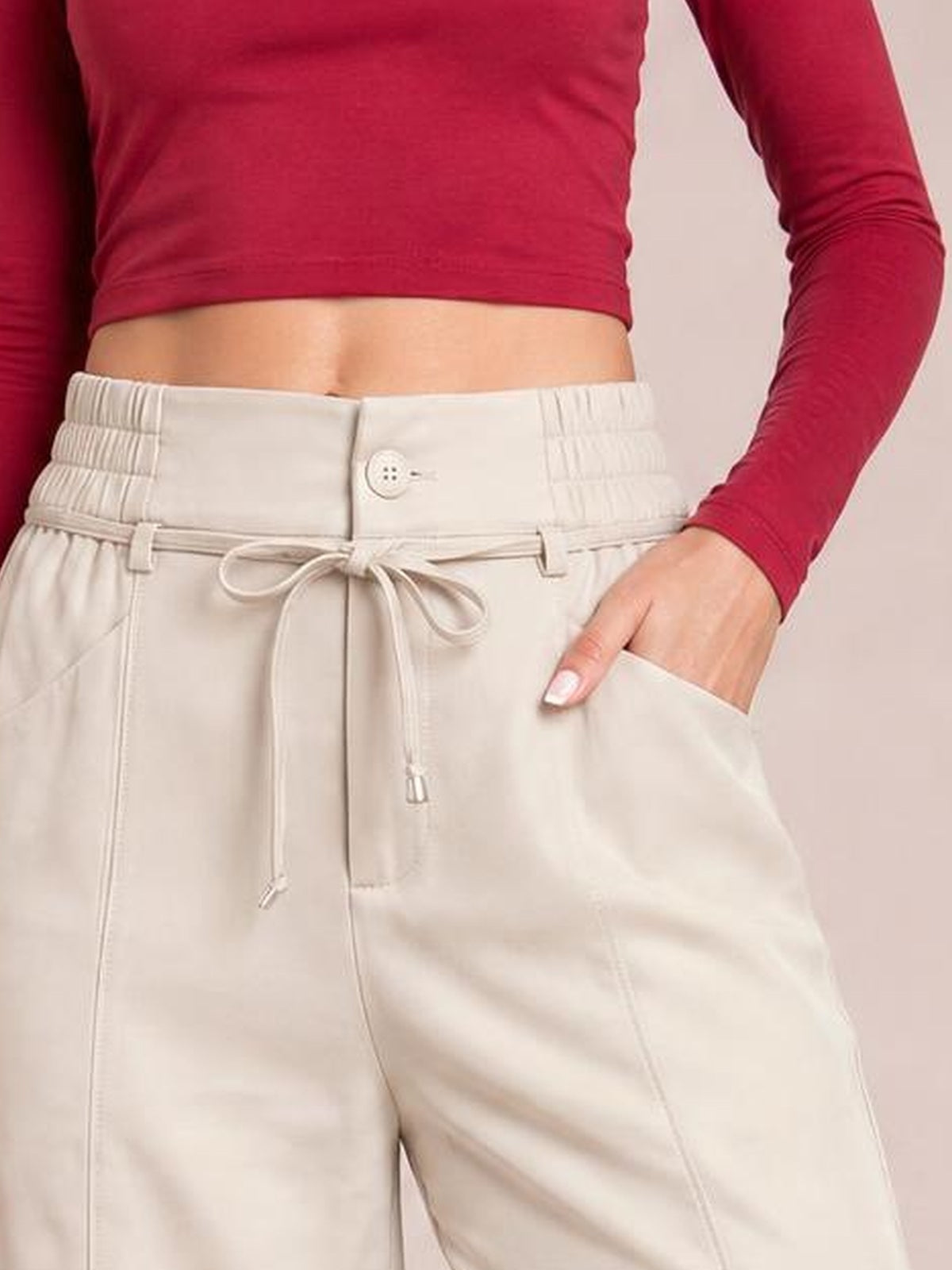 A model wears ivory high-waisted wide-leg pants with an elasticized waistband, button closure, tied drawstring, and side pocket, with one hand tucked inside.
