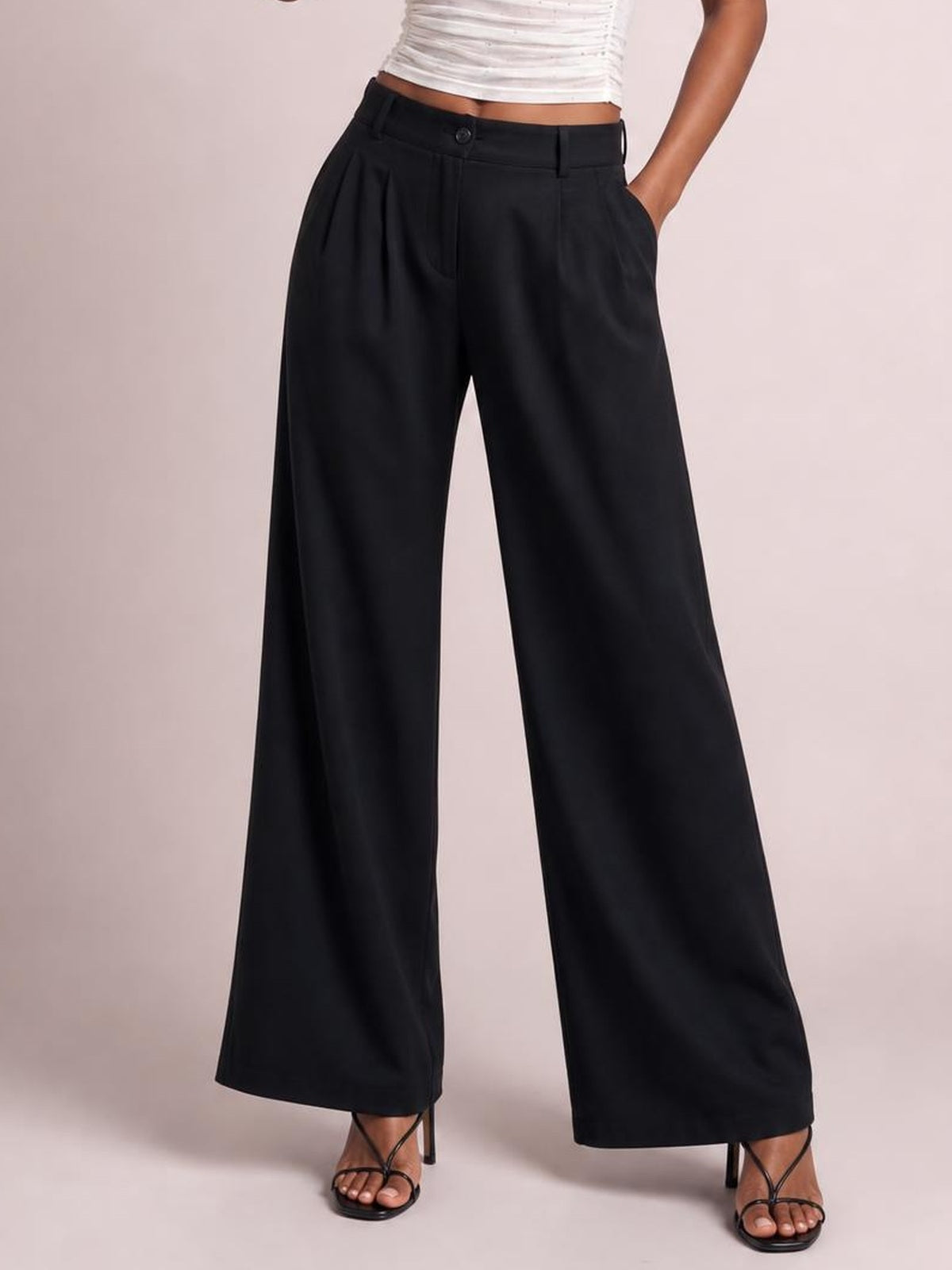 A cropped model view shows black high-waisted wide-leg pants with a button closure, belt loops, front pleats, and side pockets, worn with one hand tucked in.