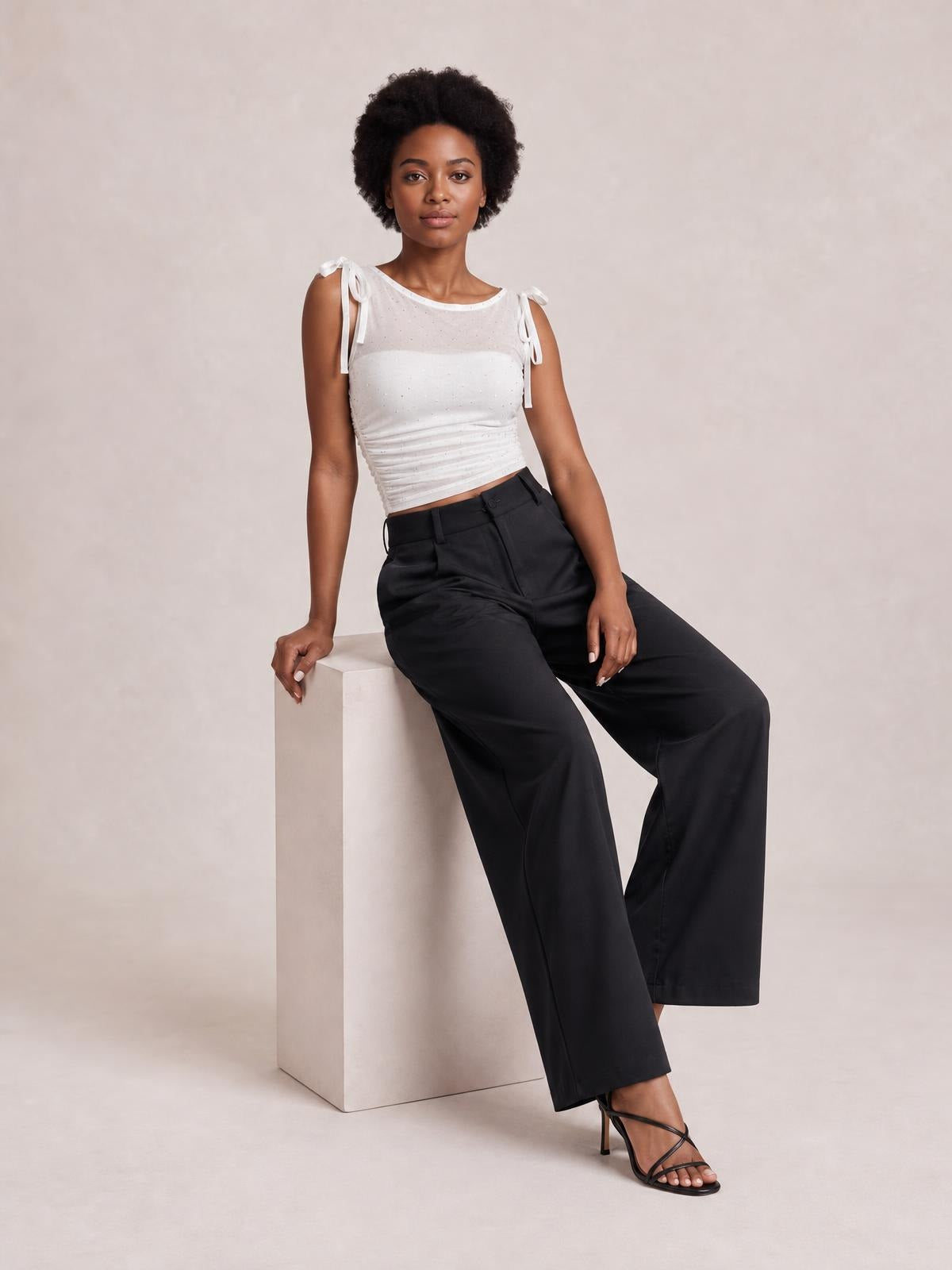 Model seated and leaning on a cube wears black high-waisted wide-leg pants with a button closure, belt loops, pockets, and a relaxed silhouette.