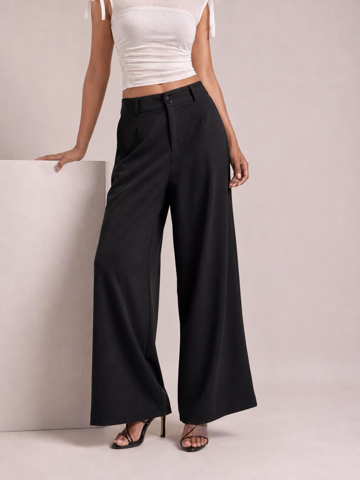 A model poses in black high-waisted wide-leg pants with a structured waistband, button closure, front pockets, and a flowing full-length silhouette.