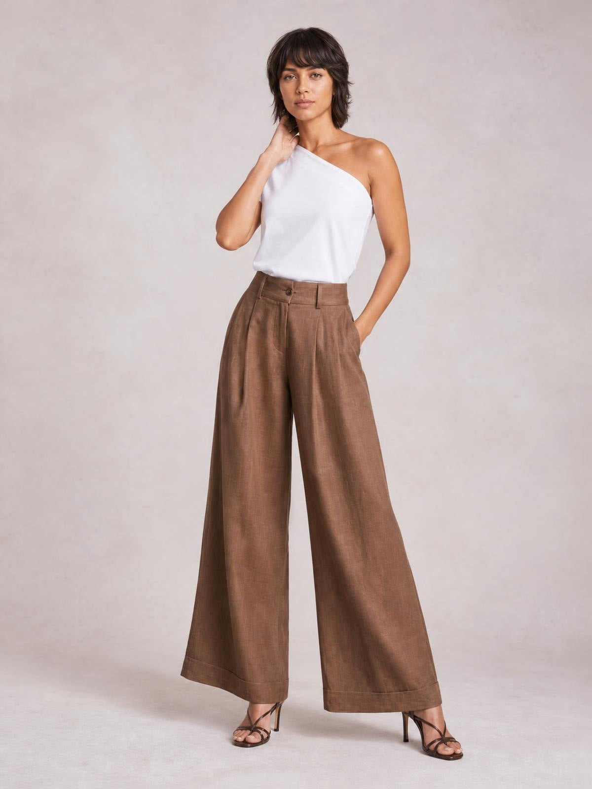 Model poses in brown high-waisted wide-leg pants with front pleats, button closure, pockets, and cuffed hems.