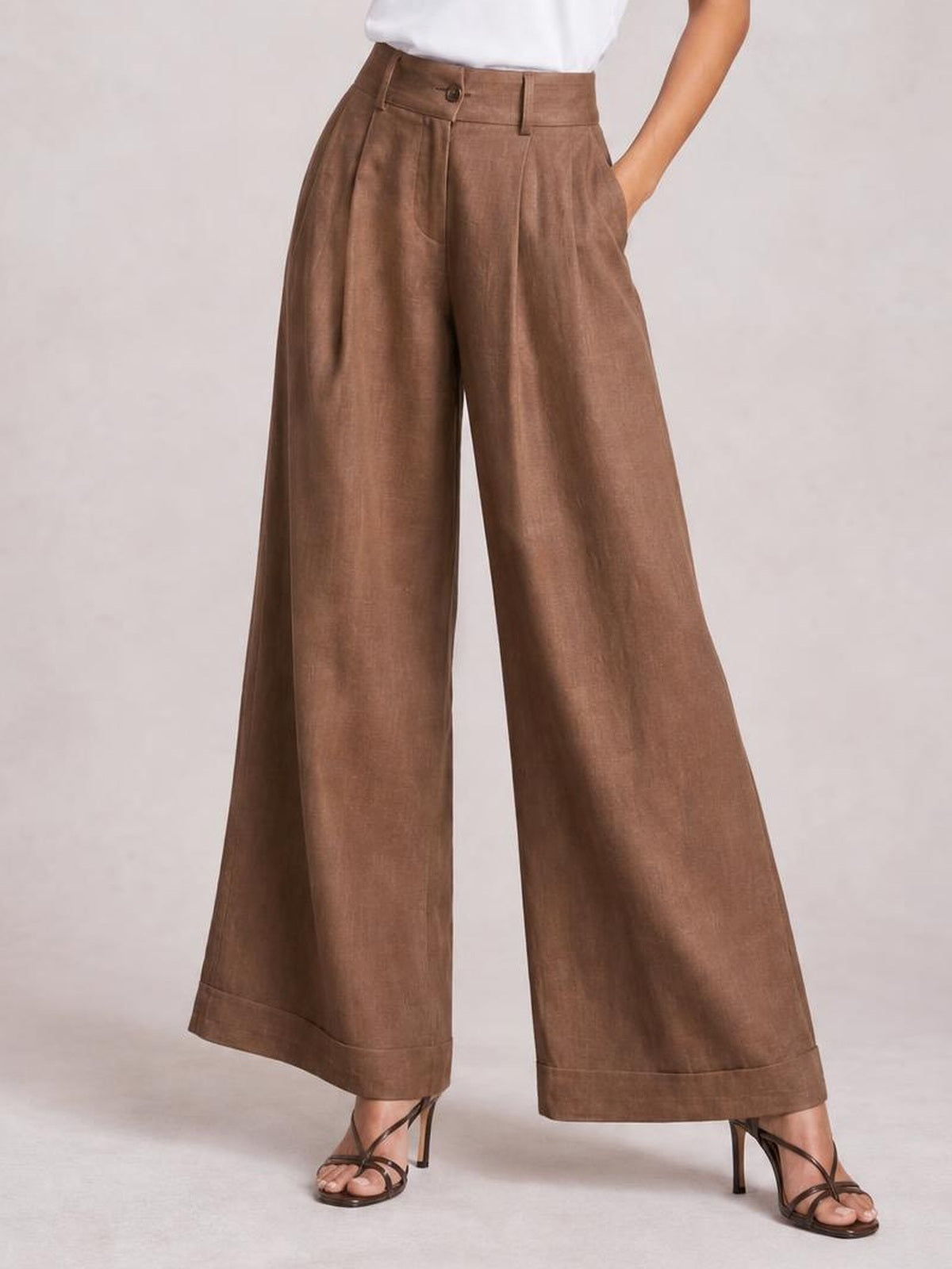 Model wearing brown high-waisted wide-leg pants with front pleats, button closure, belt loops, side pockets, and wide cuffed hems.