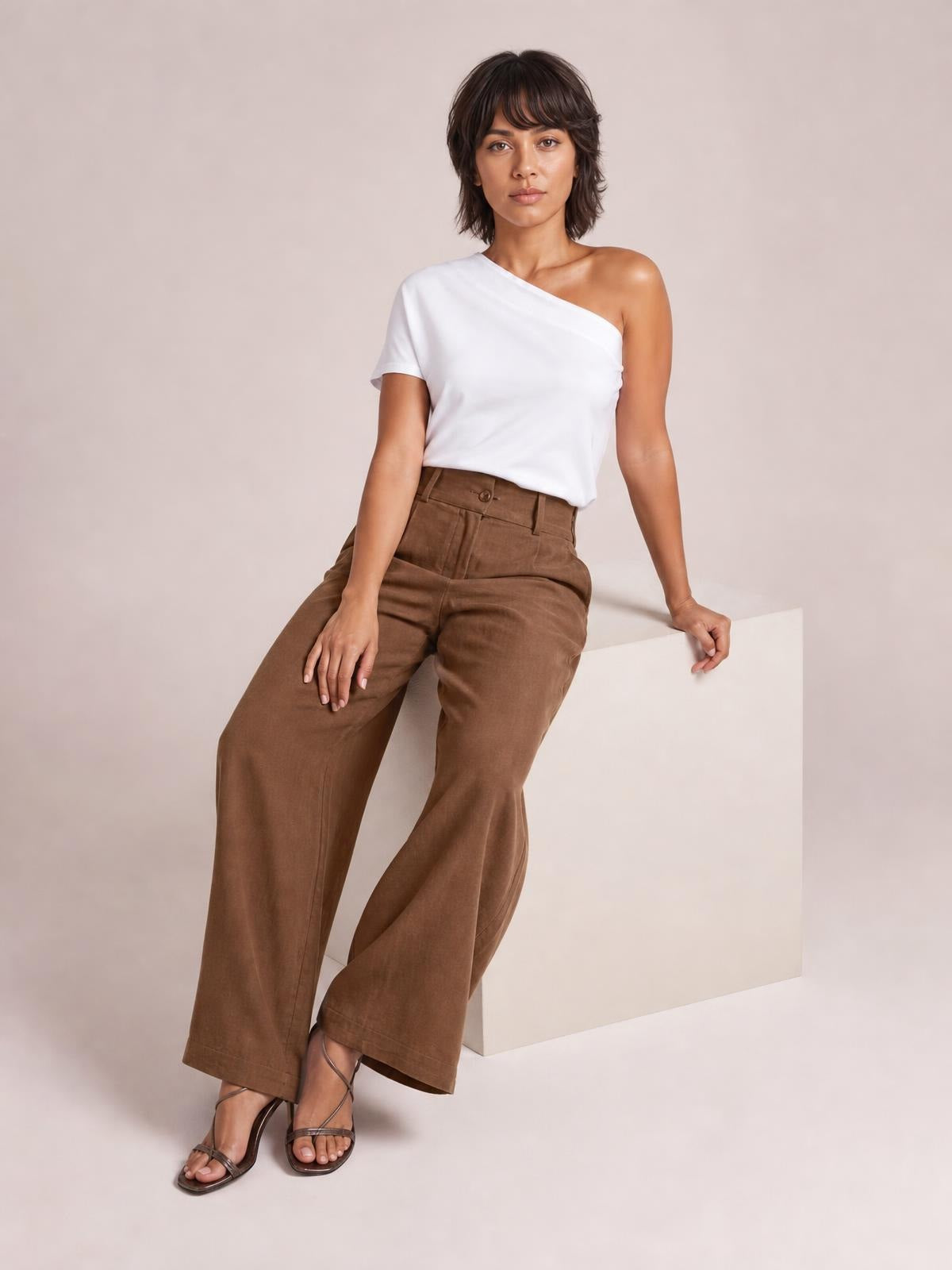 A model sits with one hand on her thigh, wearing brown high-waisted wide-leg pants with a buttoned waistband, pockets, and tailored front seams.