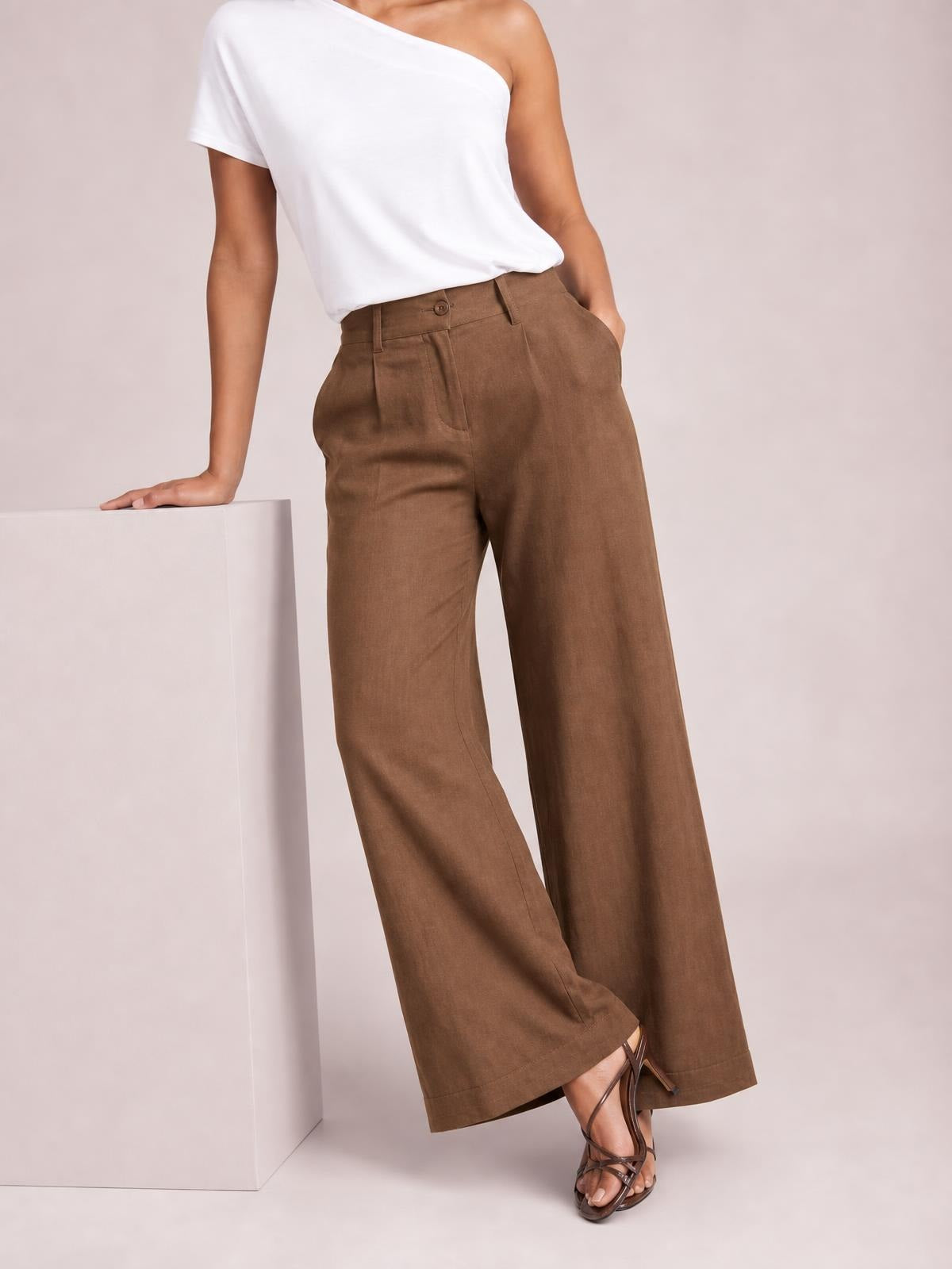 Model poses beside a pedestal in brown high-waisted wide-leg pants with a buttoned waistband, front pockets, and a relaxed flowing silhouette.