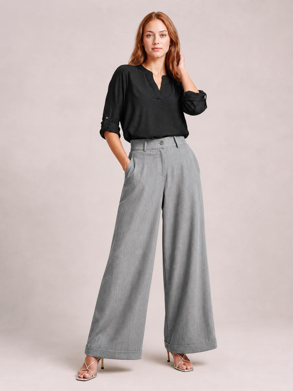 A model poses in light gray high-waisted wide-leg pants with a buttoned waistband, side pockets, tailored pleats, and cuffed hems.