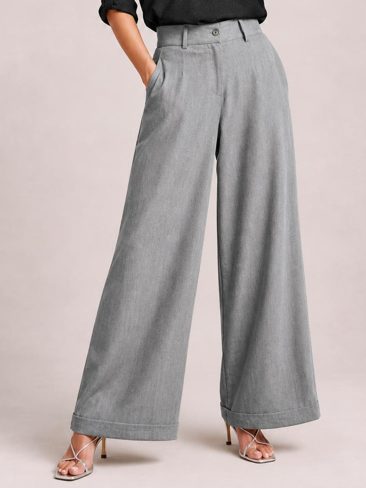 Model wears gray high-waisted wide-leg pants with a buttoned waistband, belt loops, slant pockets, and cuffed hems while standing in strappy heels.