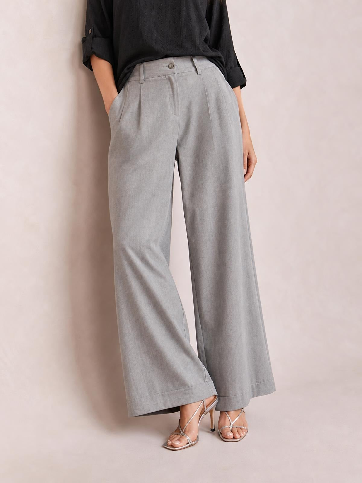 Model poses with hands in the pockets of light gray high-waisted wide-leg pants featuring a button closure, belt loops, front pleats, and cuffed hems.