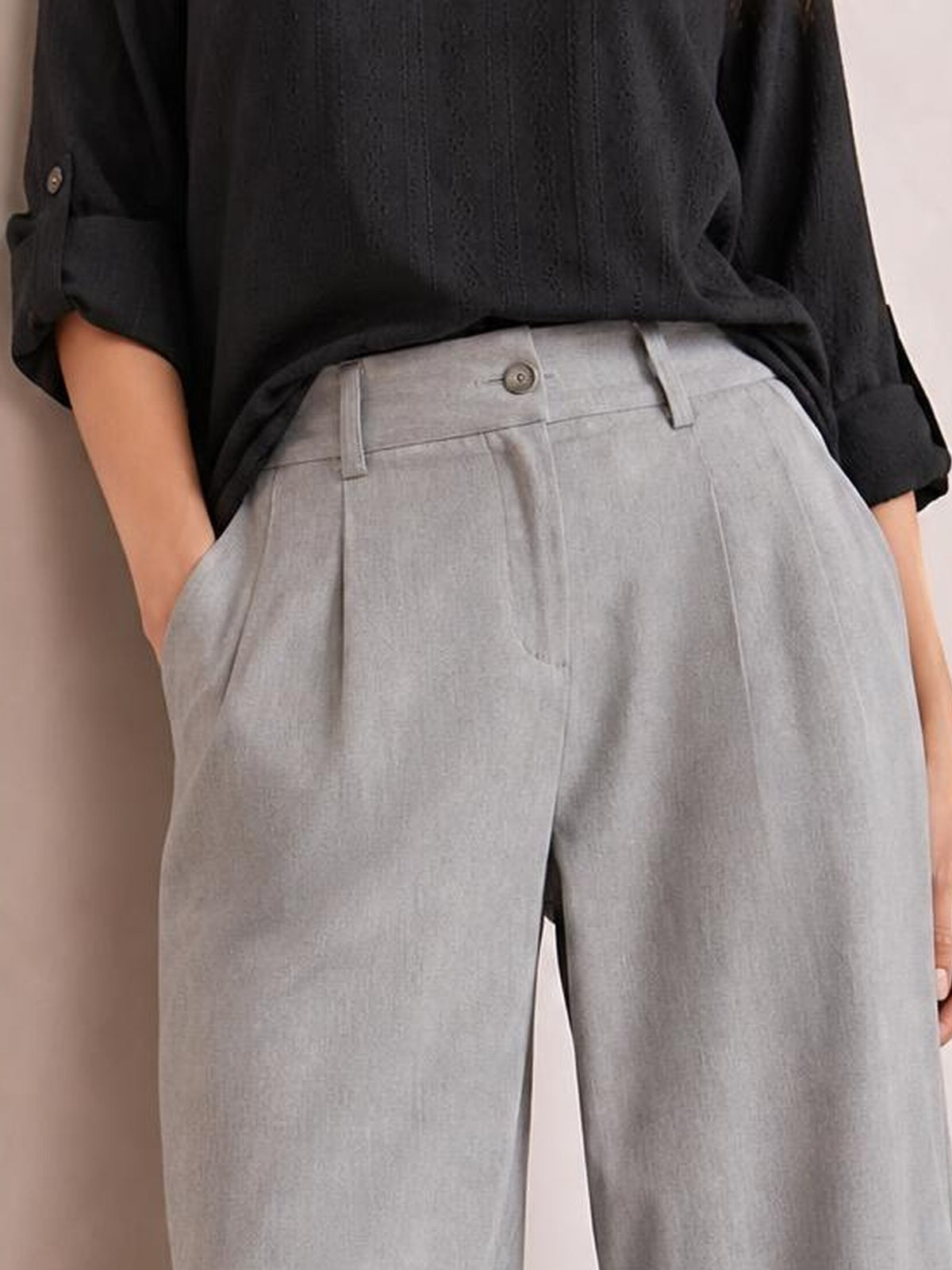 A model wears light gray high-waisted wide-leg pants with a buttoned waistband, belt loops, front pockets, and a relaxed silhouette, hands tucked in pockets.