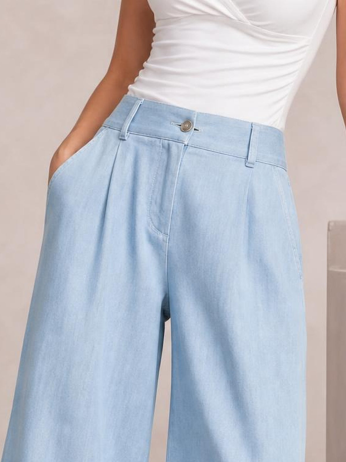 A model wears light blue high-waisted wide-leg pants with a button closure, belt loops, front pockets, and a relaxed silhouette, posing with hands in the pockets.