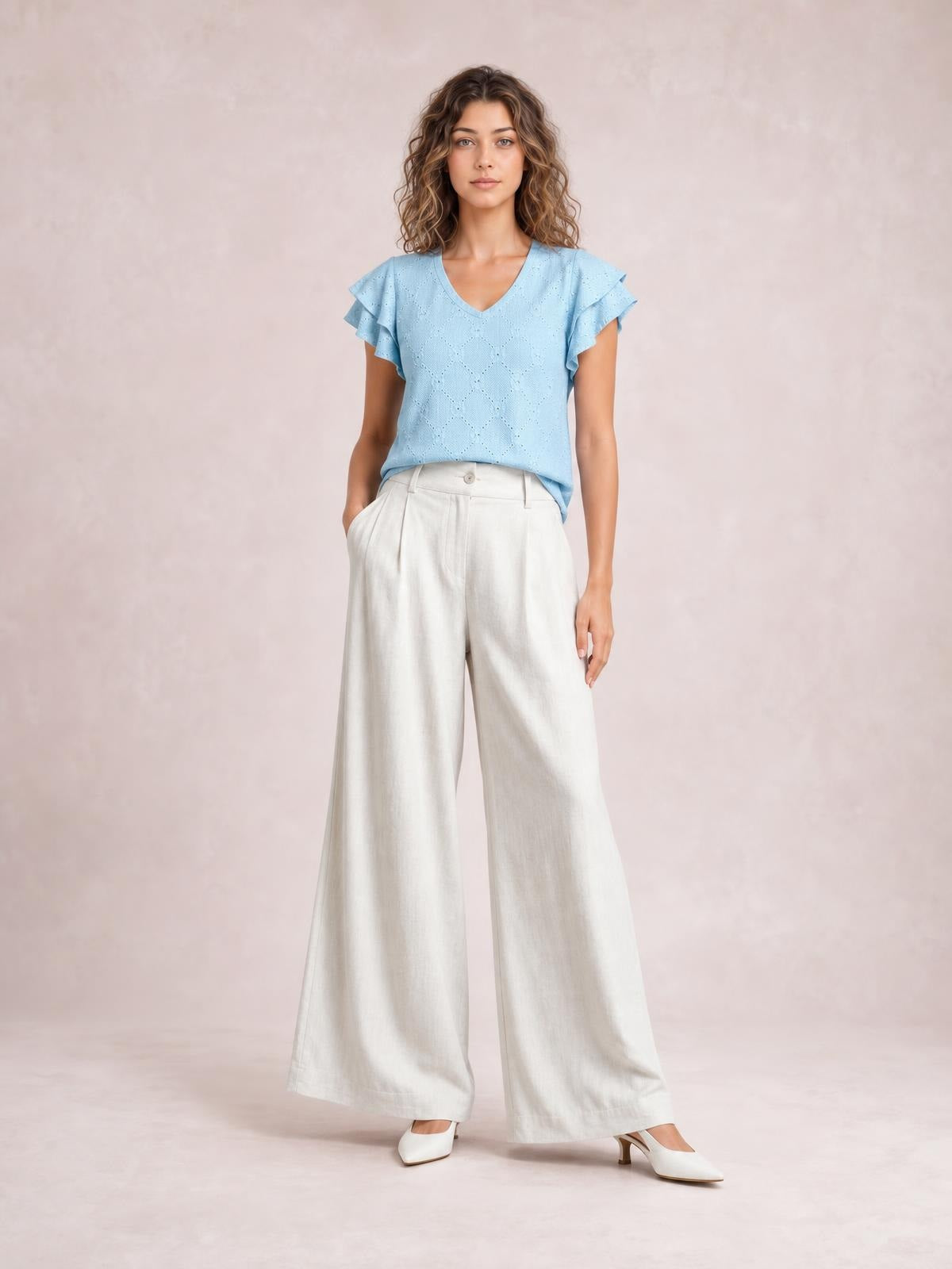 A model stands with one hand in a pocket, wearing high-waisted ivory wide-leg pants with a button closure, front pleats, and a flowing full-length silhouette.