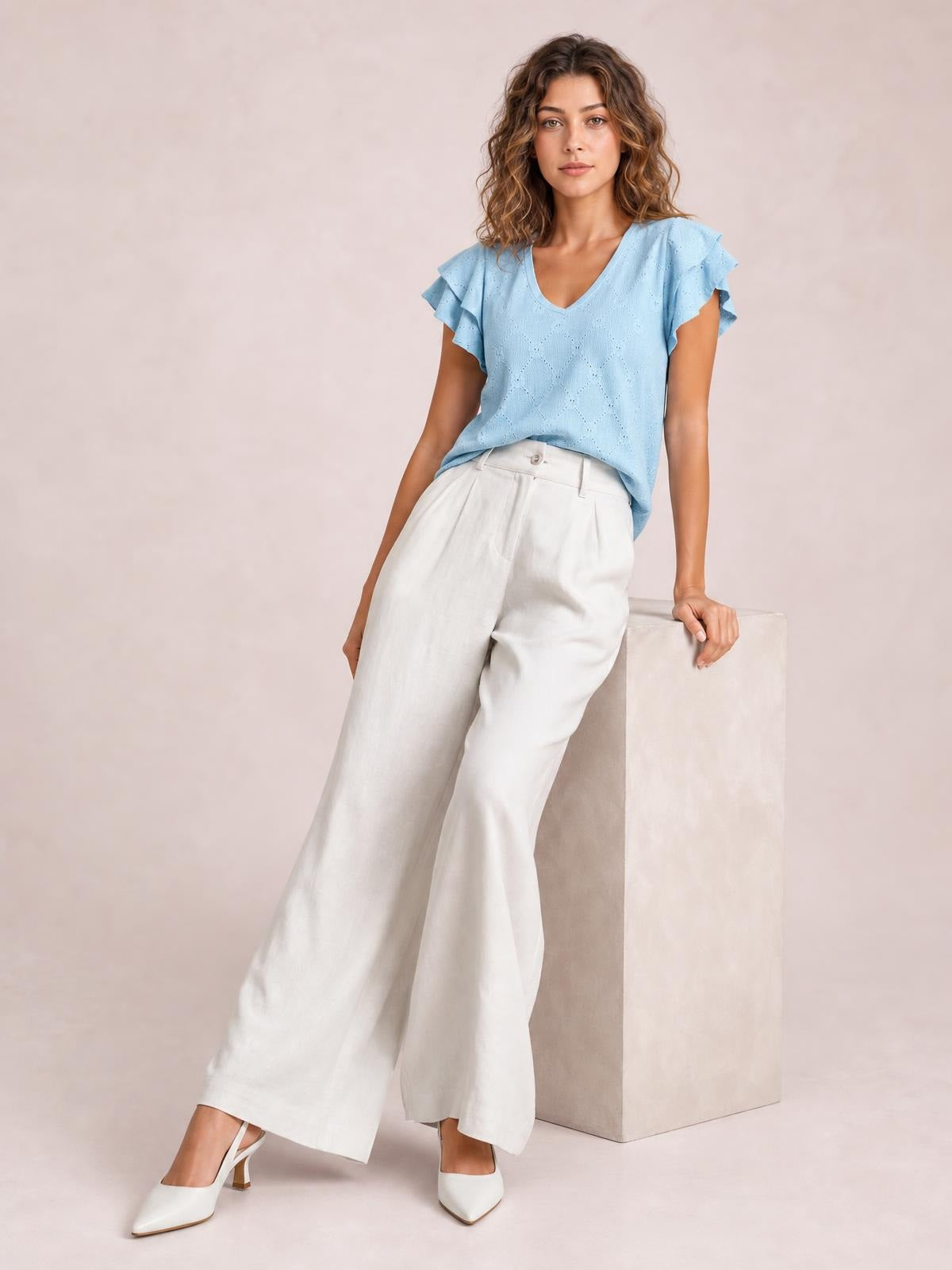 Model poses in white high-waisted wide-leg pants with a buttoned waistband, front pleats, and relaxed flowing silhouette.