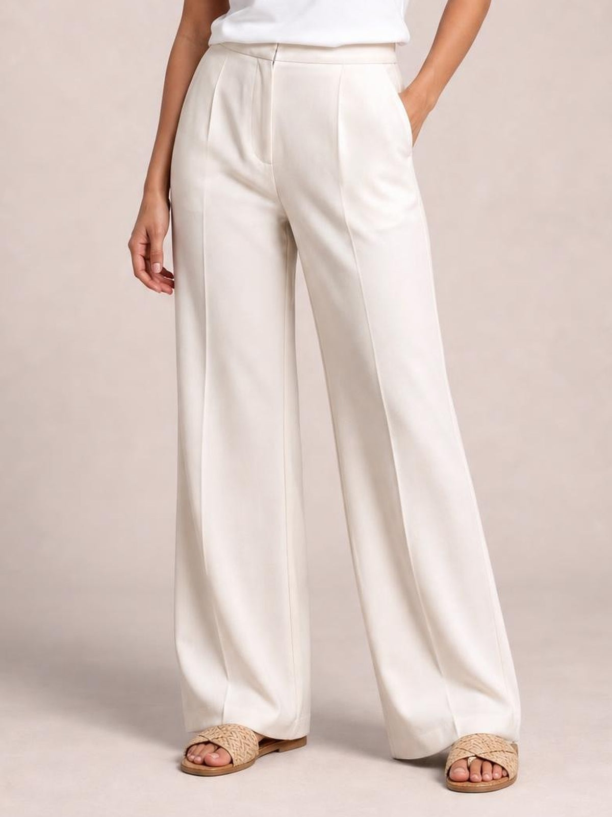 Model wears high-waisted white wide-leg pants with a structured waistband, front pleats, vertical seams, and side pockets, posing with one hand tucked in.