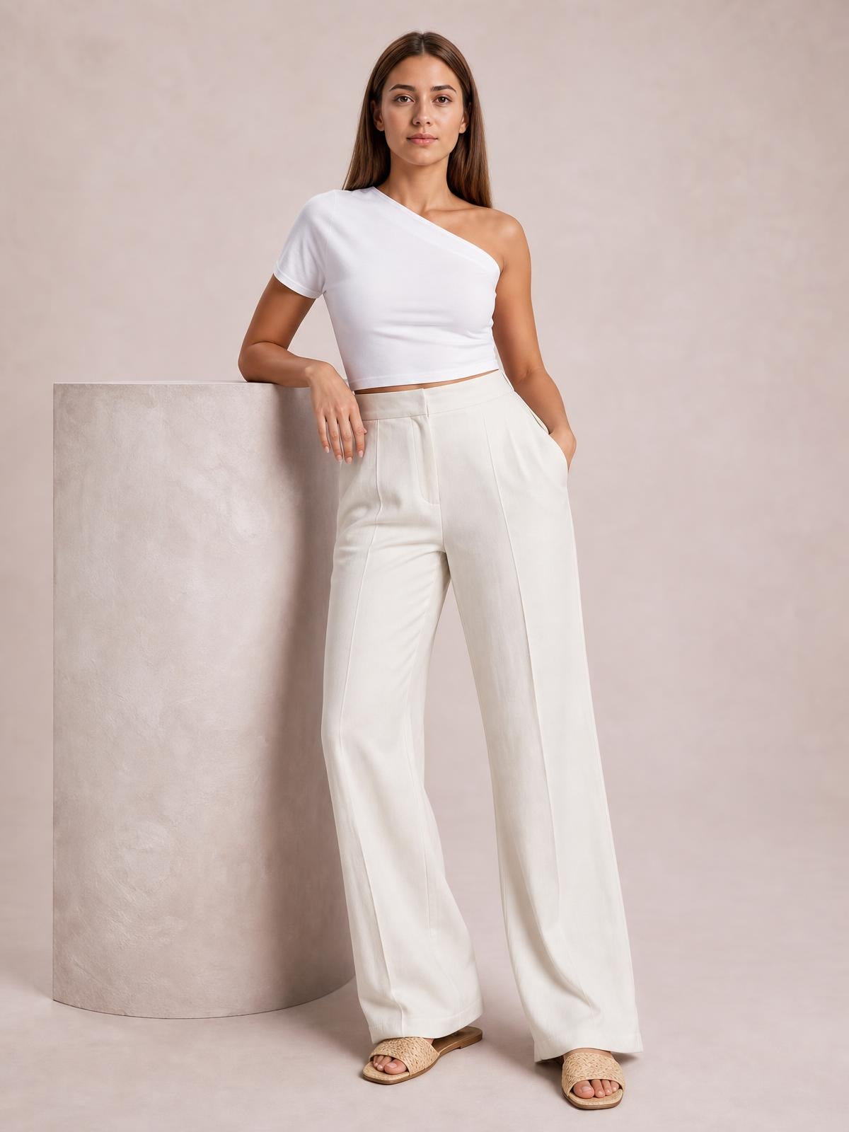 A model leans against a column wearing white high-waisted wide-leg pants with front pleats, side pockets, and a relaxed full-length silhouette.
