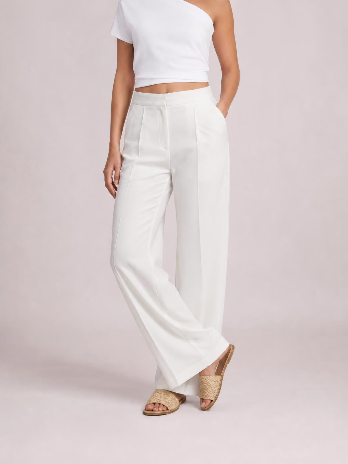 A model wears white high-waisted wide-leg pants with front pleats and side pockets, standing with one hand in a pocket and ankles crossed.
