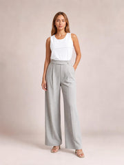 A model stands in light gray high-waisted wide-leg pleated pants with a structured waistband and side pockets, posing with one hand tucked in a pocket.