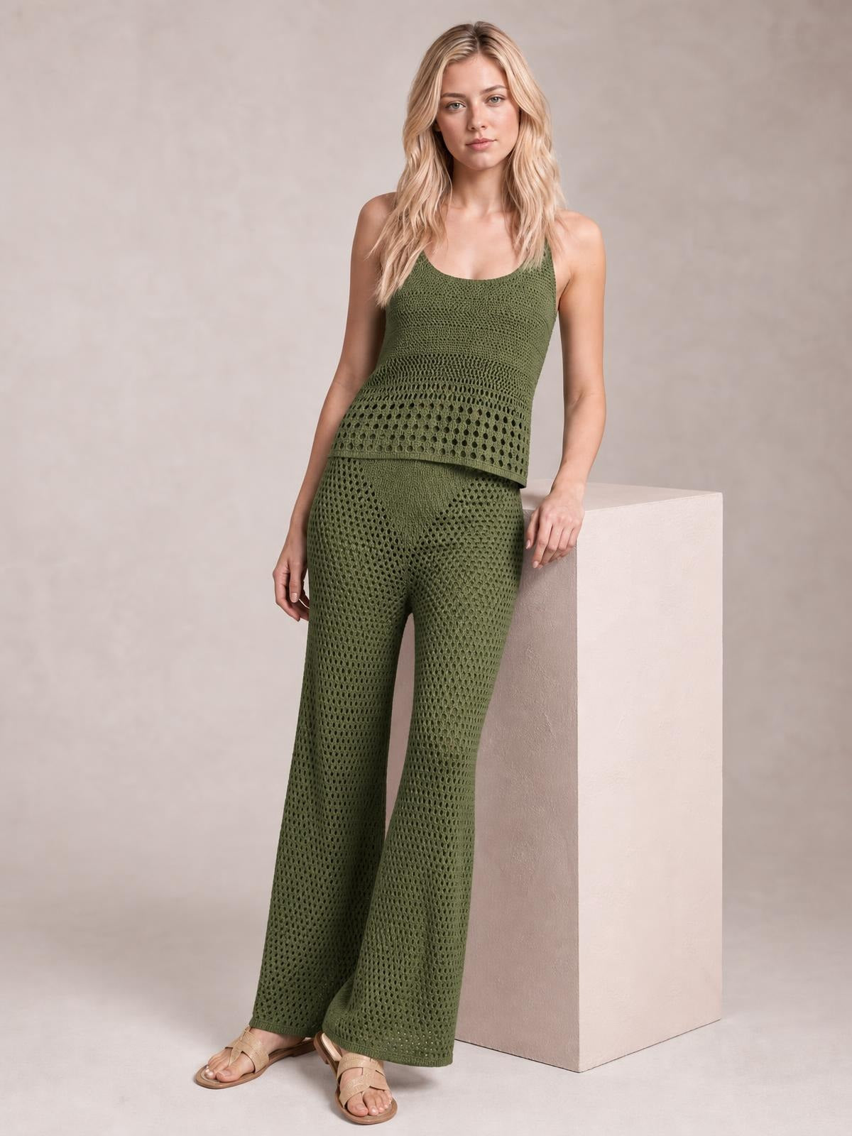 A model wears an olive-green hollow-out knit top with a layered hem and matching wide-leg pants, leaning casually with one hand on a pedestal.