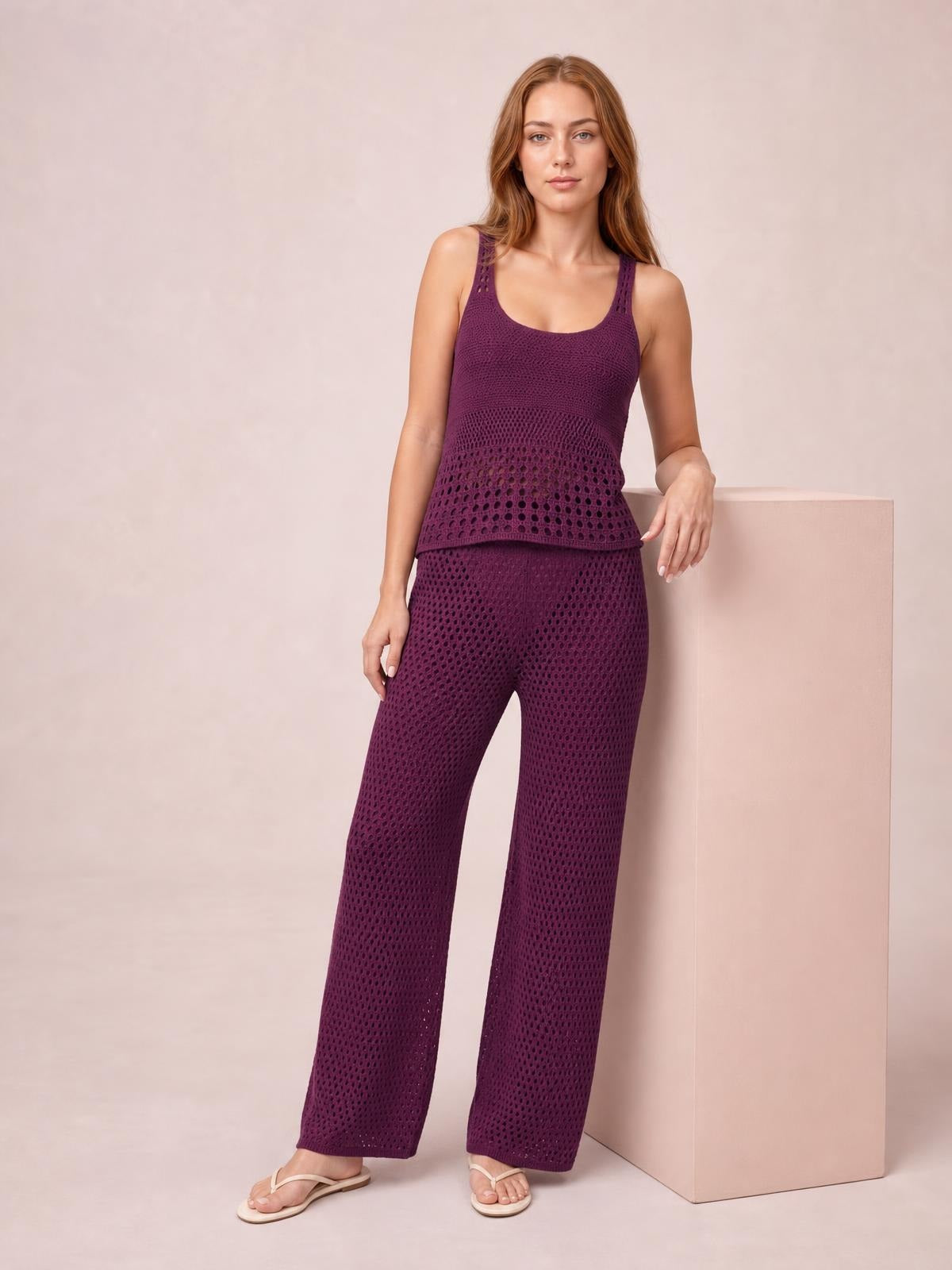 A model wears a plum-purple hollow-out round-neck knit top with openwork panels and matching wide-leg pants, leaning casually with one hand on a box.