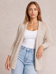 Model wears a cream hollowed knit open-front cardigan with openwork stitching, long sleeves, ribbed cuffs, and a relaxed longline silhouette over a white tank top.