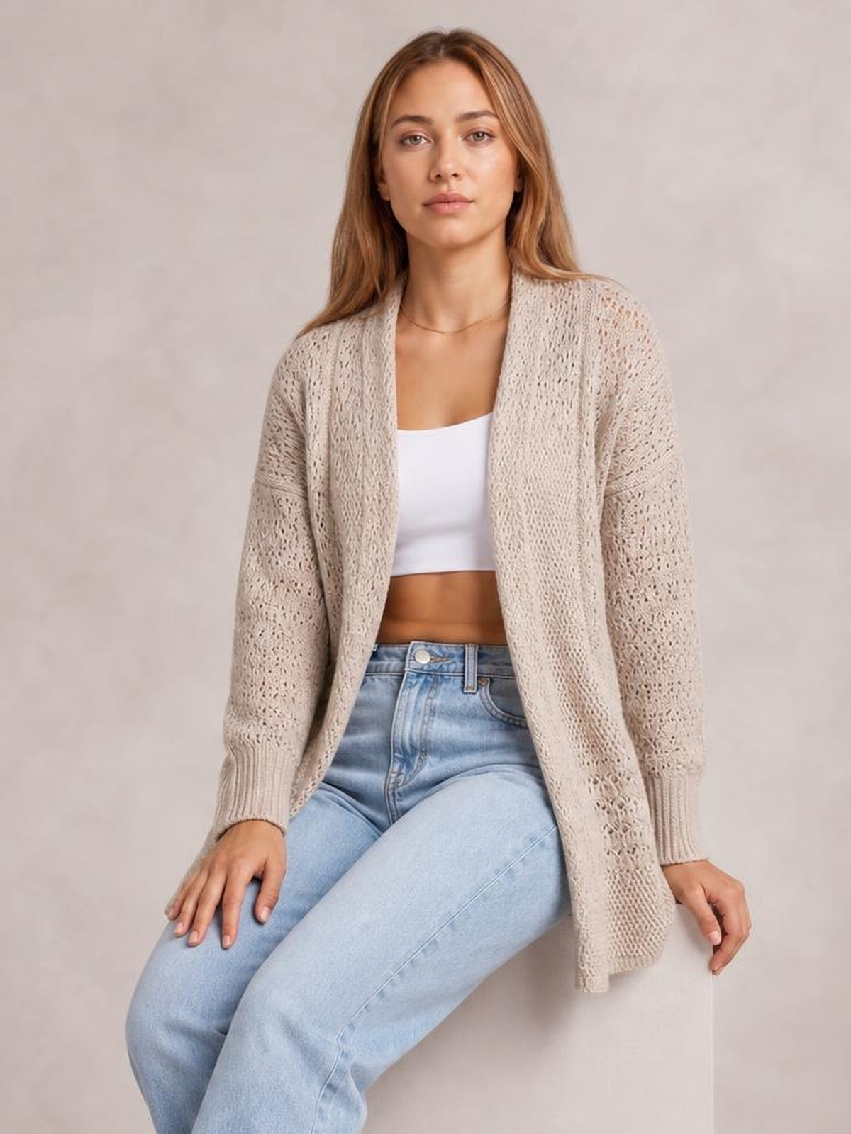 A seated model wears a beige hollowed knit open-front cardigan with an airy openwork texture, long sleeves, ribbed cuffs, and a relaxed silhouette.