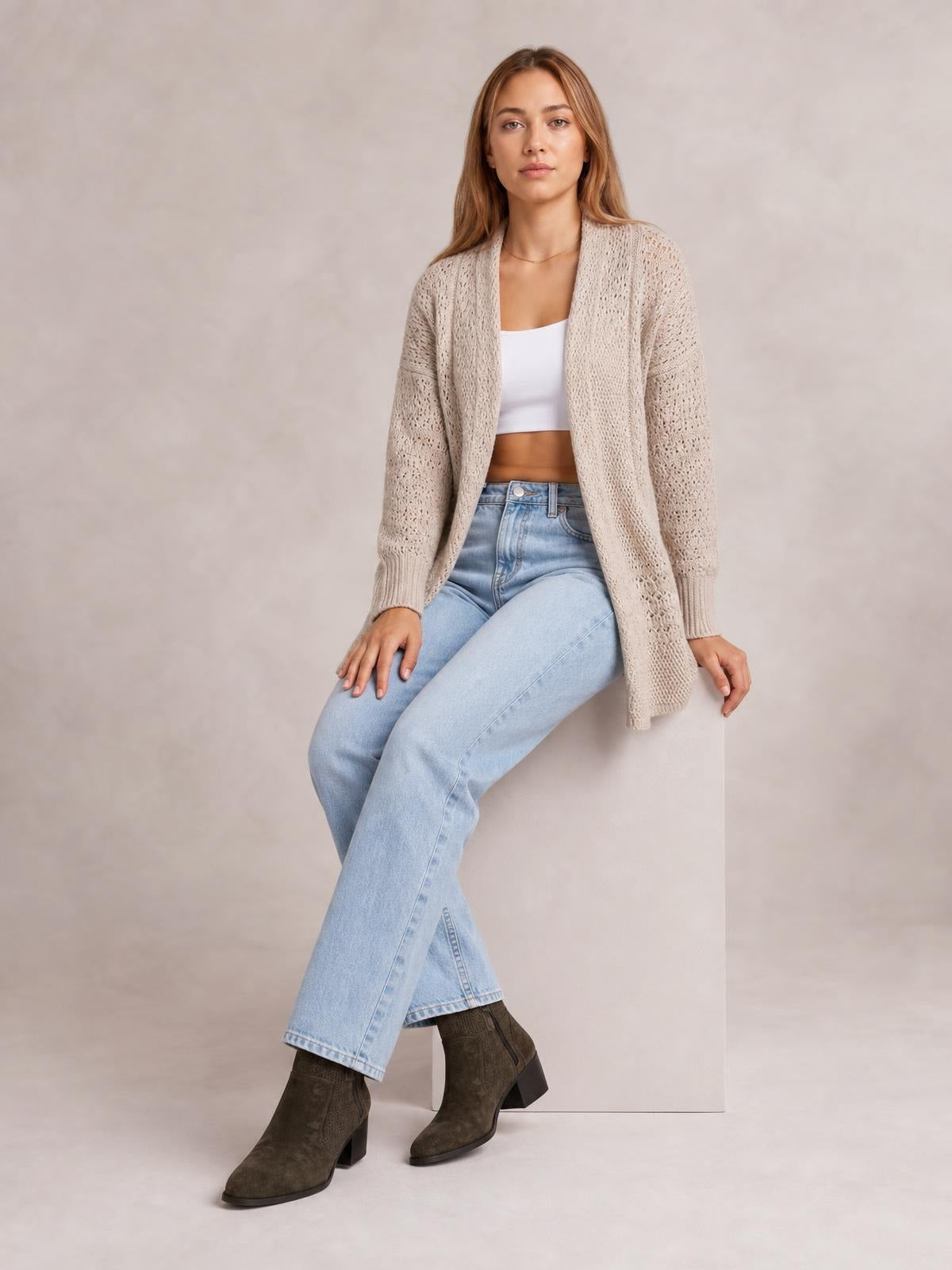 Model seated in a beige open-front cardigan with hollowed openwork knit, long sleeves, ribbed cuffs, and a relaxed longline silhouette.