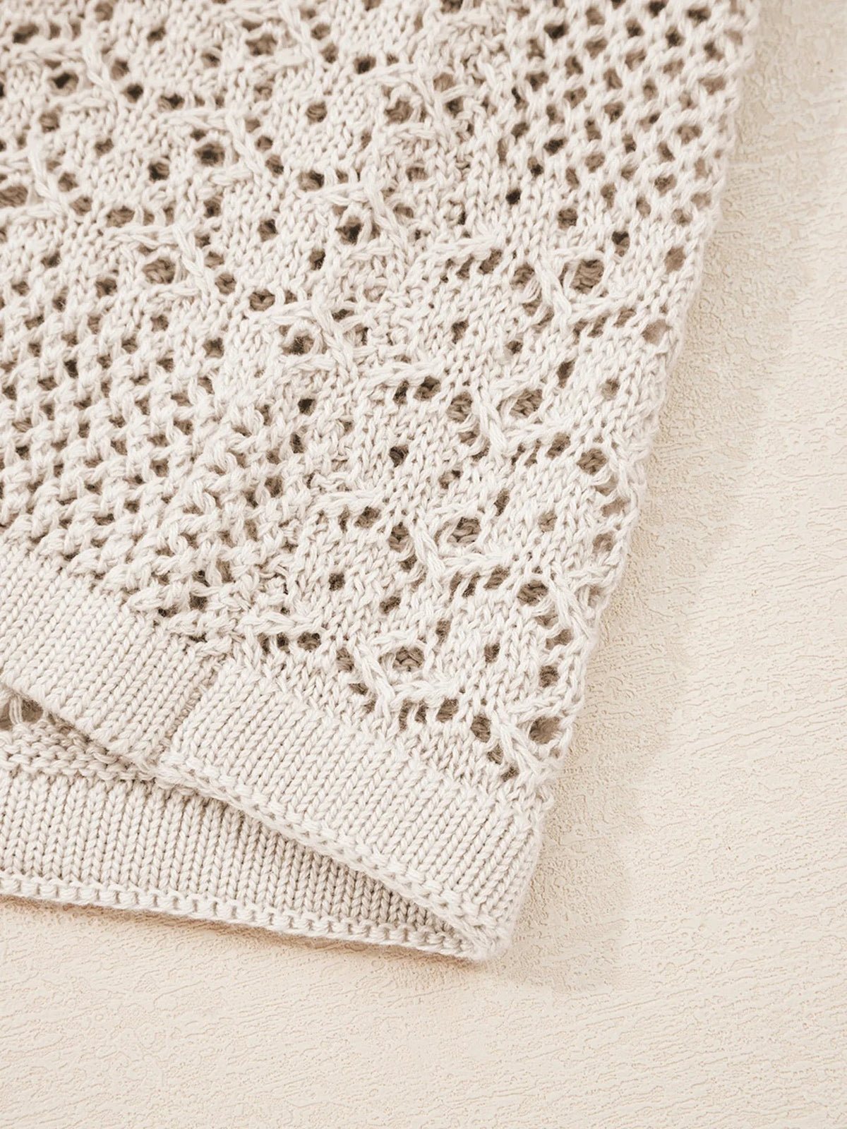 Cream openwork knit cardigan fabric shown close up, featuring a breathable hollowed texture and ribbed trim along the garment edge.