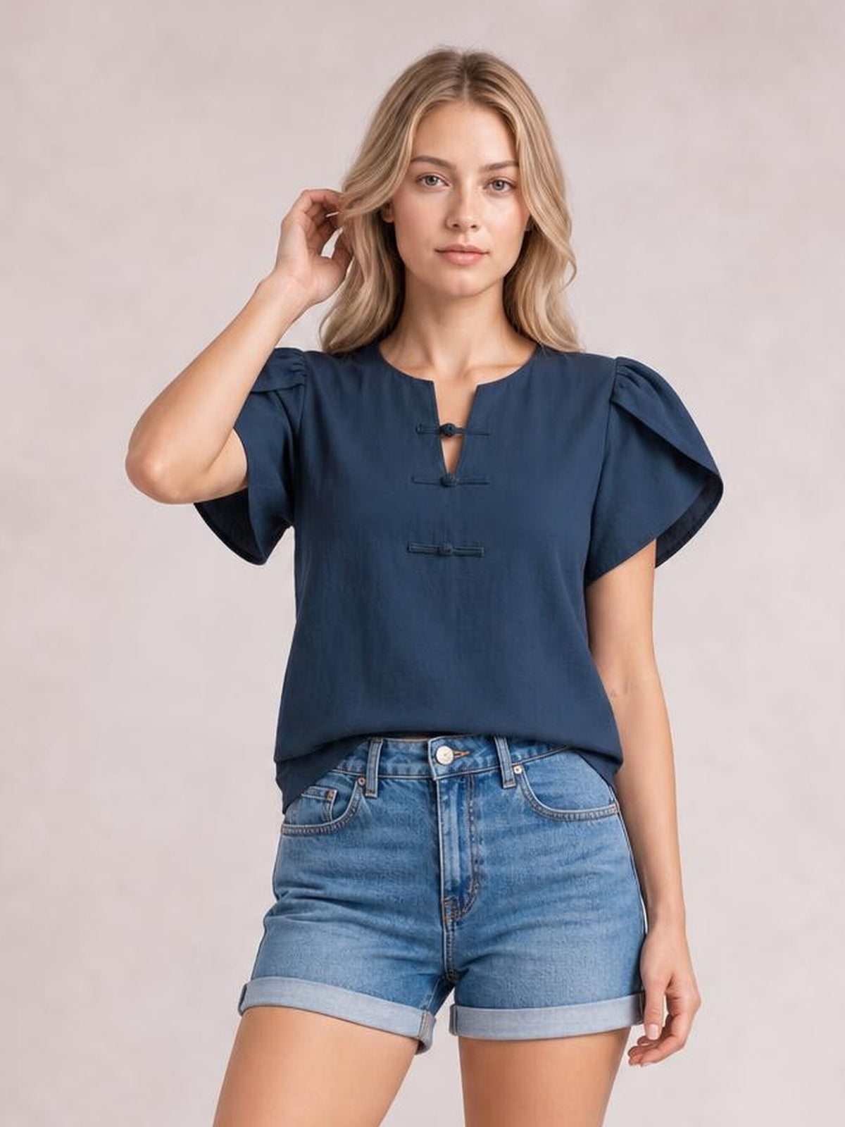 Model wearing a navy notched short sleeve blouse with a relaxed silhouette, gathered shoulders, flutter sleeves, and loop closures while touching her hair.