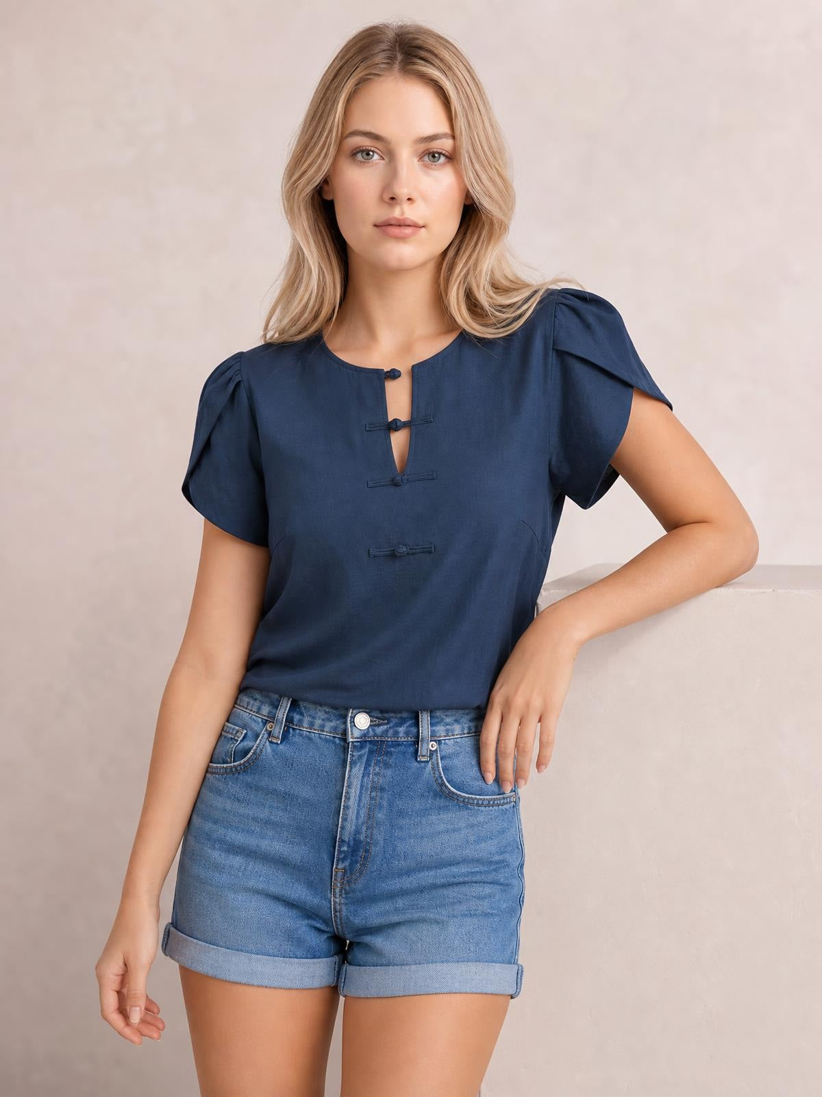 A model wears a navy notched short sleeve blouse with a relaxed straight silhouette, gathered shoulders, three front loop closures, and one hand on her hip.
