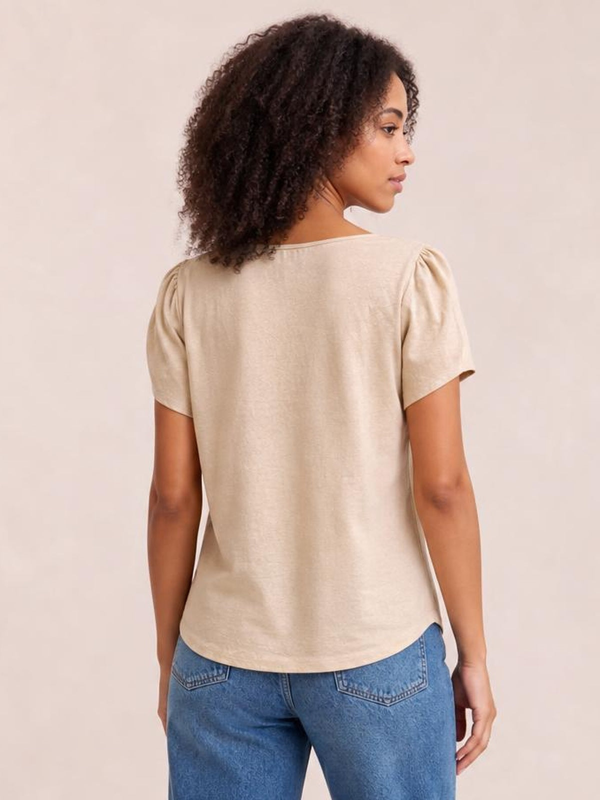 A model wears a honey-colored short sleeve blouse with gathered shoulders, a relaxed straight silhouette, and a gently curved hem, shown from the back.