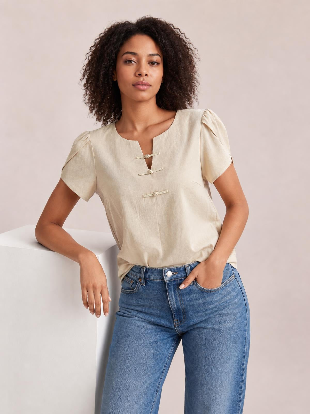A model wears a honey notched short sleeve blouse with a relaxed silhouette, rounded neckline, and three front loop closures, leaning with one hand in her jeans pocket.