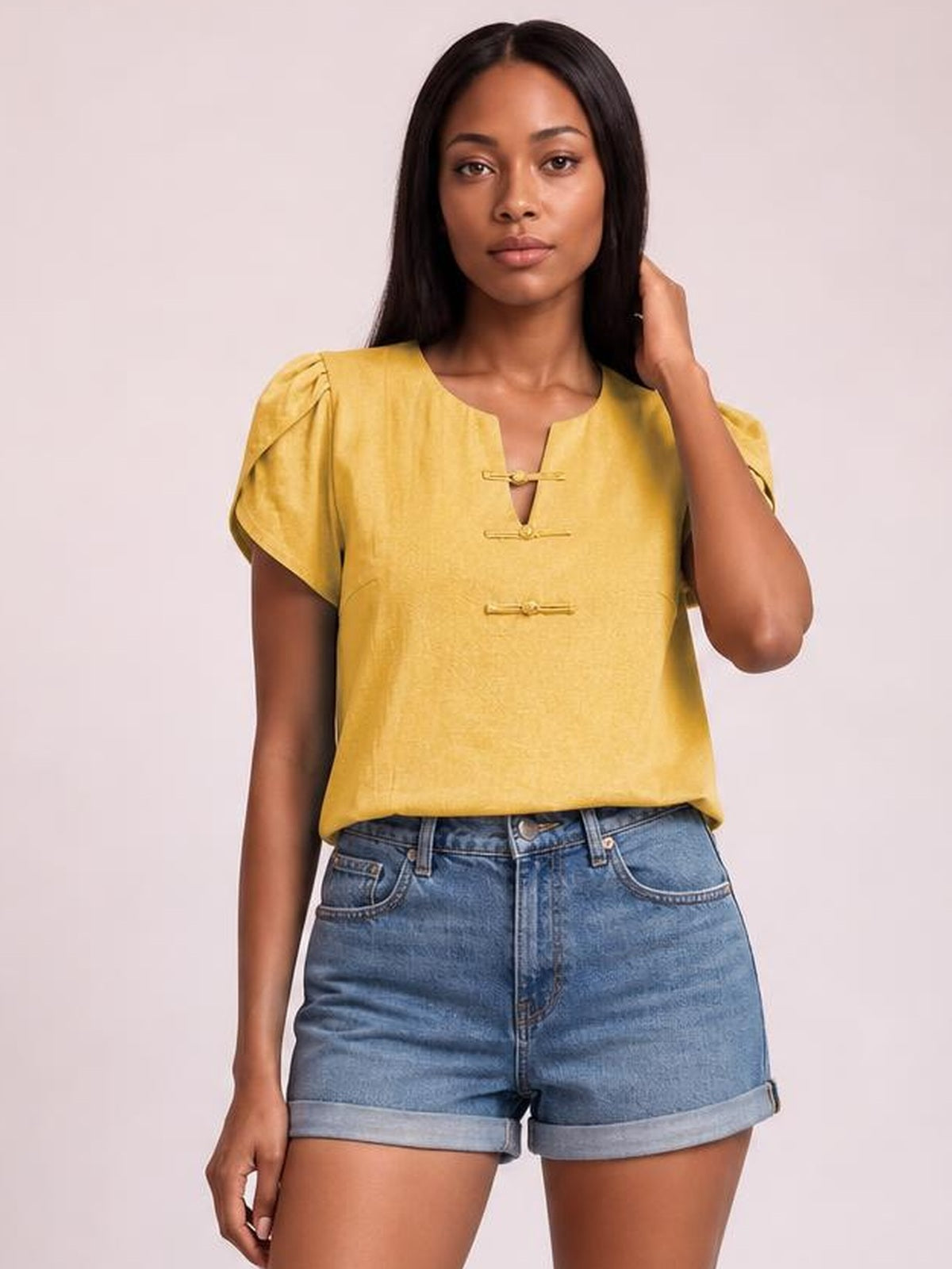 Model wearing a honey yellow notched short sleeve blouse with a relaxed silhouette, gathered shoulders, and front loop closures, posing with one hand in her hair.