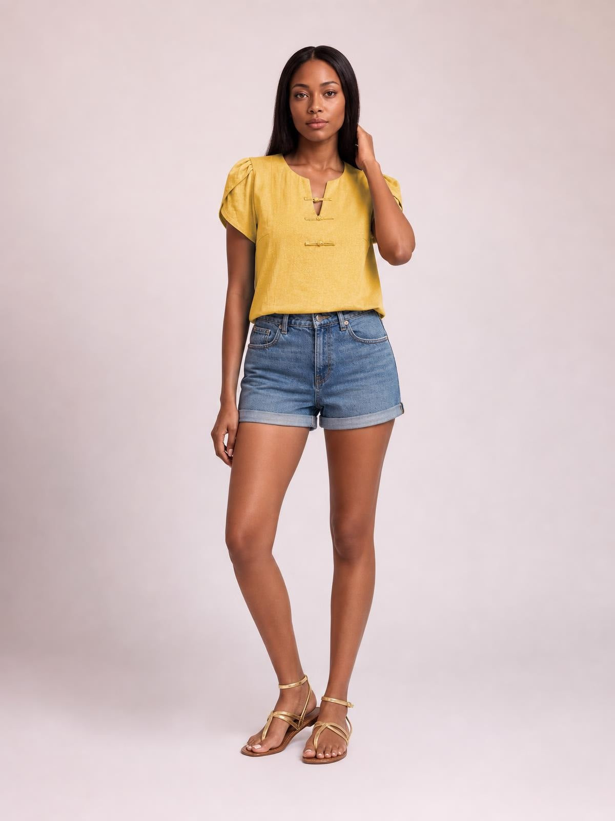 A model poses in a honey-yellow notched short sleeve blouse with gathered shoulders, a relaxed silhouette, and decorative front closures, paired with denim shorts.