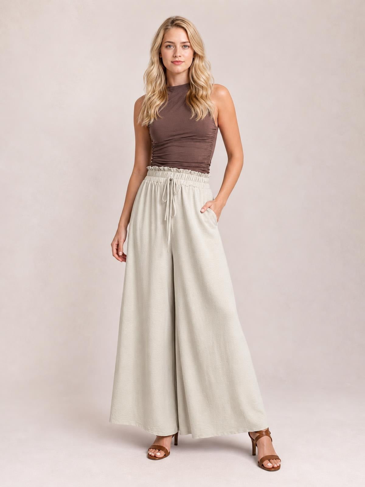 Model poses in cream tied high-waist wide-leg pants with a gathered waistband, front drawstring, and flowing full-length silhouette.