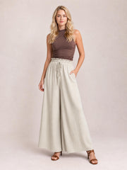Model poses in cream tied high-waist wide-leg pants with a gathered waistband, front drawstring, and flowing full-length silhouette.