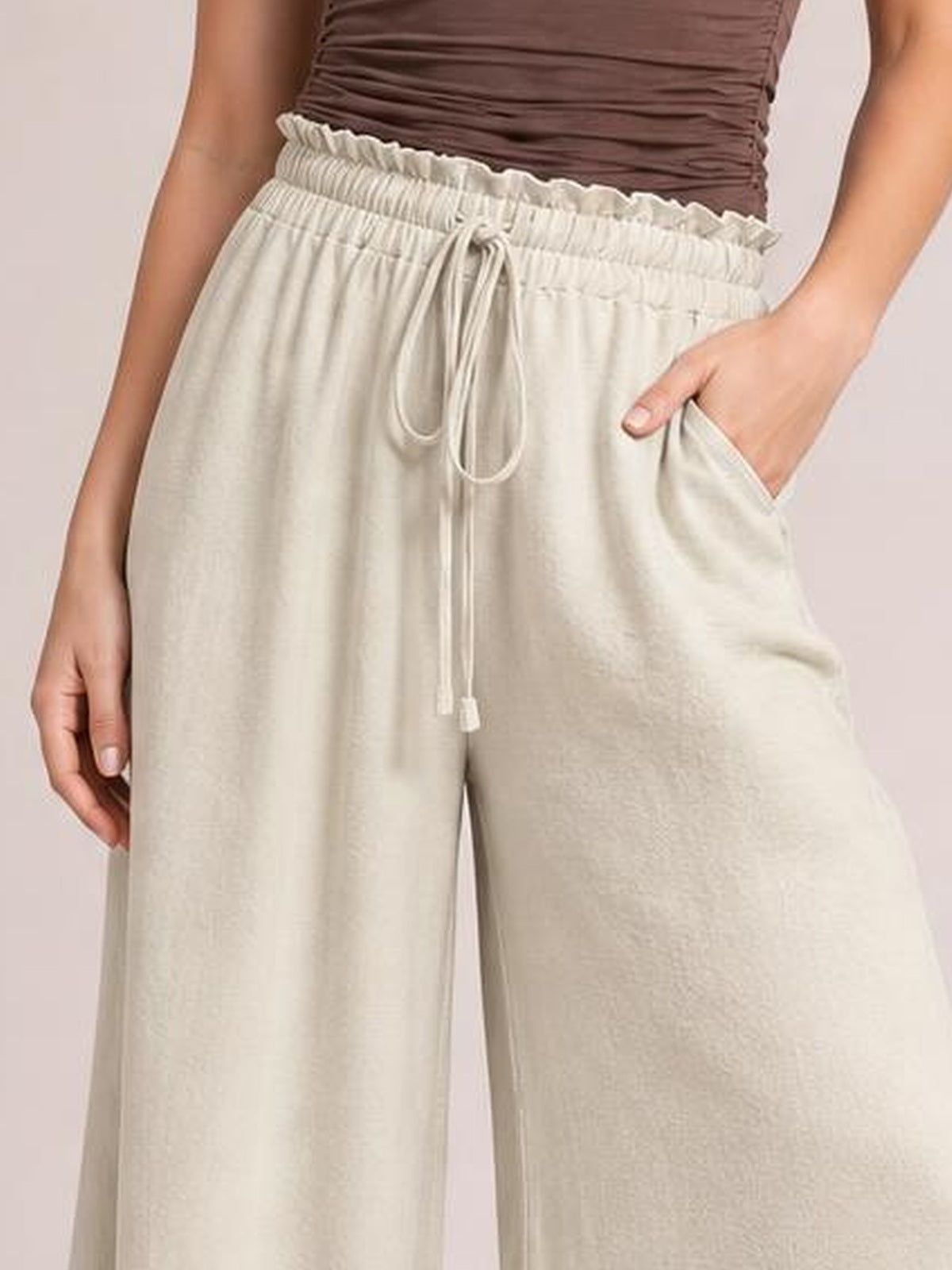 A model wears opaque cream high-waist wide-leg pants with a ruffled elastic waistband, front tie detail, and side pockets, posing with one hand in a pocket.