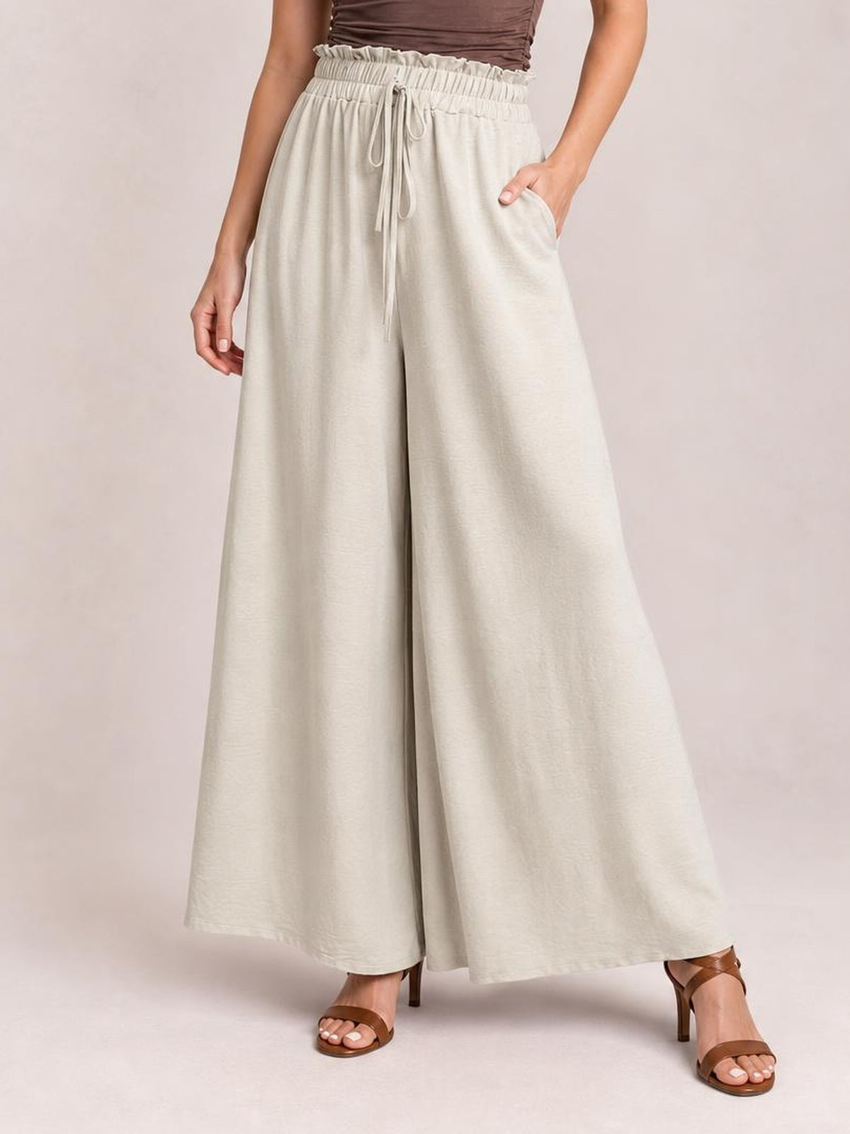 Model wearing light beige tied high waist wide leg pants with a gathered waistband, drawstring, side pockets, and flowing full-length silhouette.