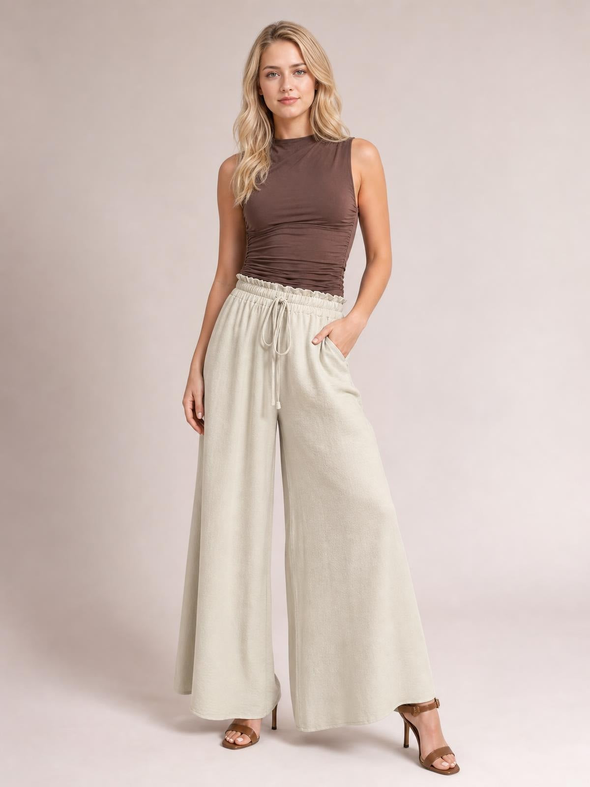 A model poses in cream tied high-waist wide-leg pants with an elastic waistband, drawstring front, side pockets, and a flowing full-length silhouette.