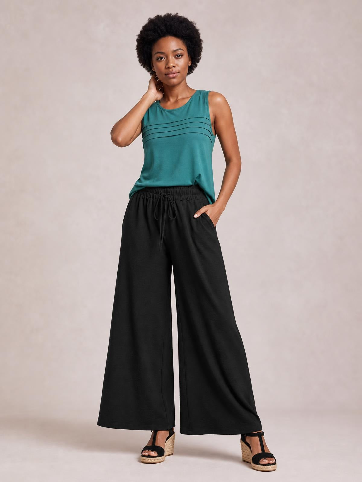 Model wearing black tied high waist wide leg pants with an elasticized drawstring waist and flowing full-length silhouette, posing with one hand in a pocket.