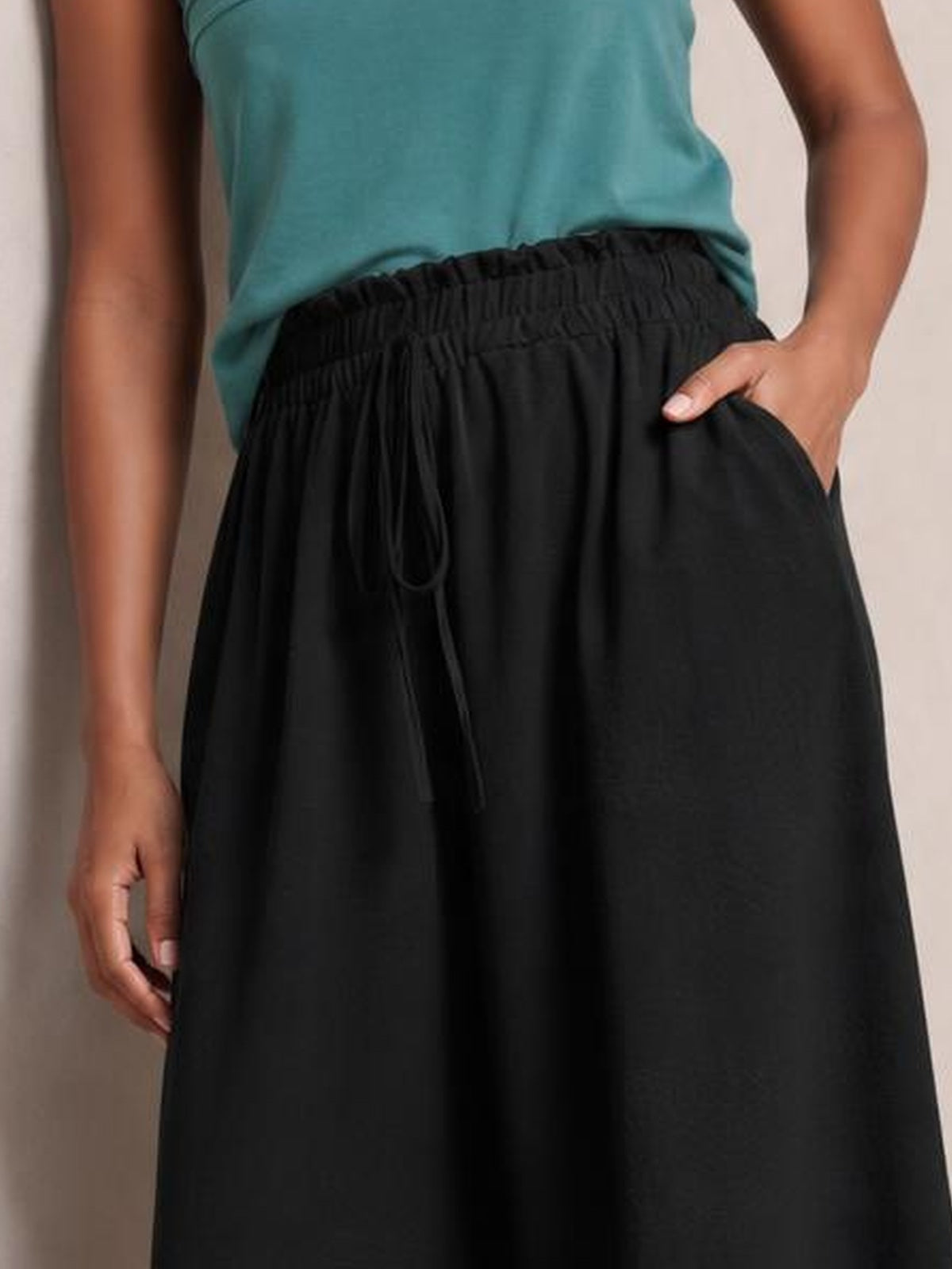Model wearing black tied high-waist wide-leg pants with an elastic waistband, drawstring front, and relaxed flowing silhouette, with one hand in a pocket.