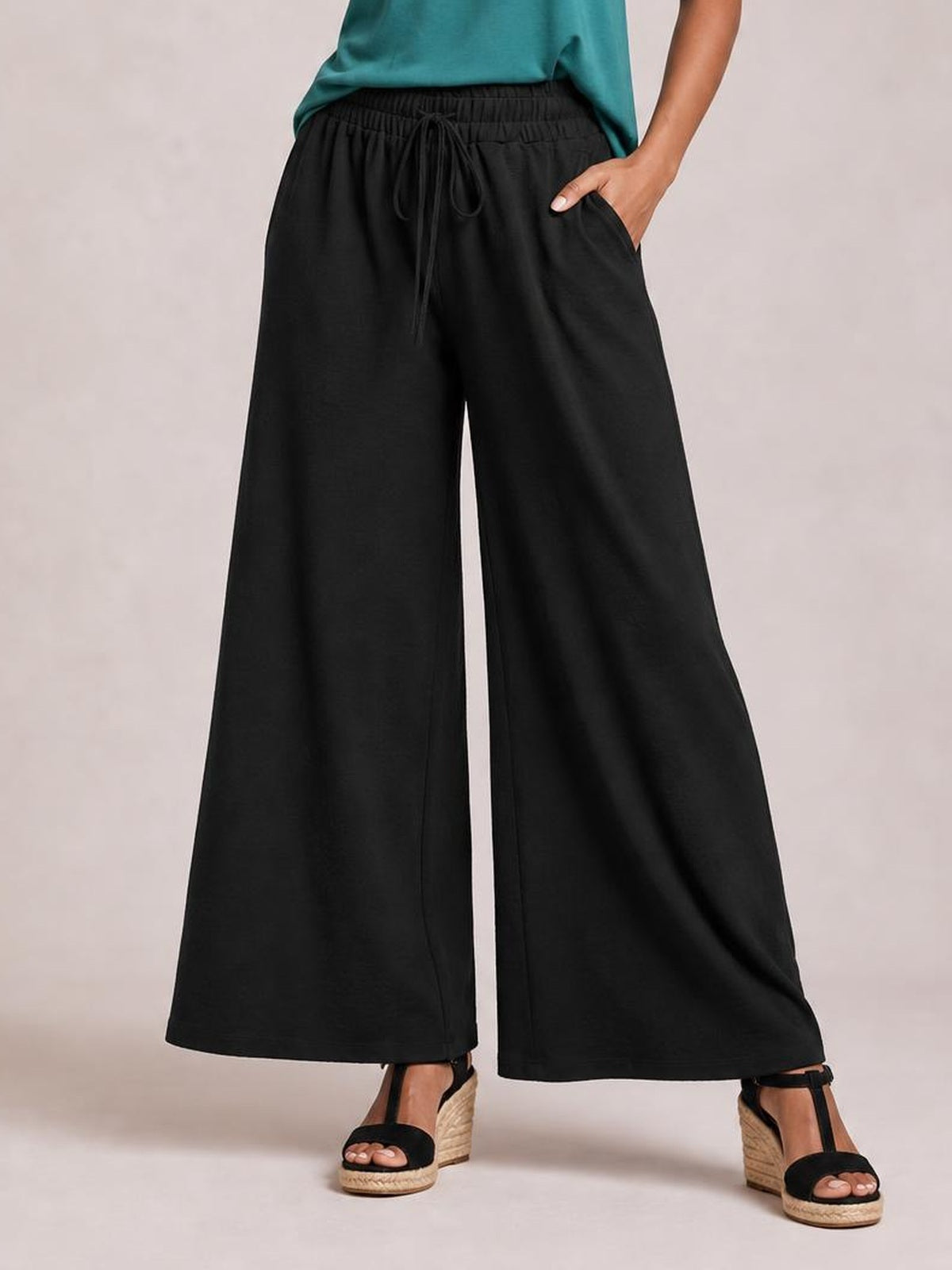 A model wears black tied high-waist wide-leg pants with an elastic drawstring waistband, flowing silhouette, side pockets, and one hand tucked inside.