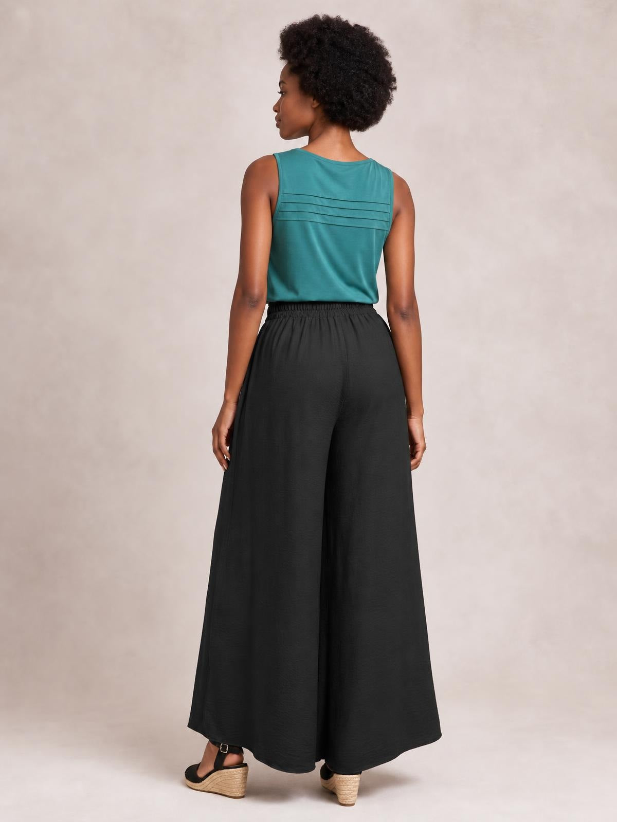 Model shown from the back wearing black tied high waist wide leg pants with a flowing, full-length silhouette and gathered waistband.