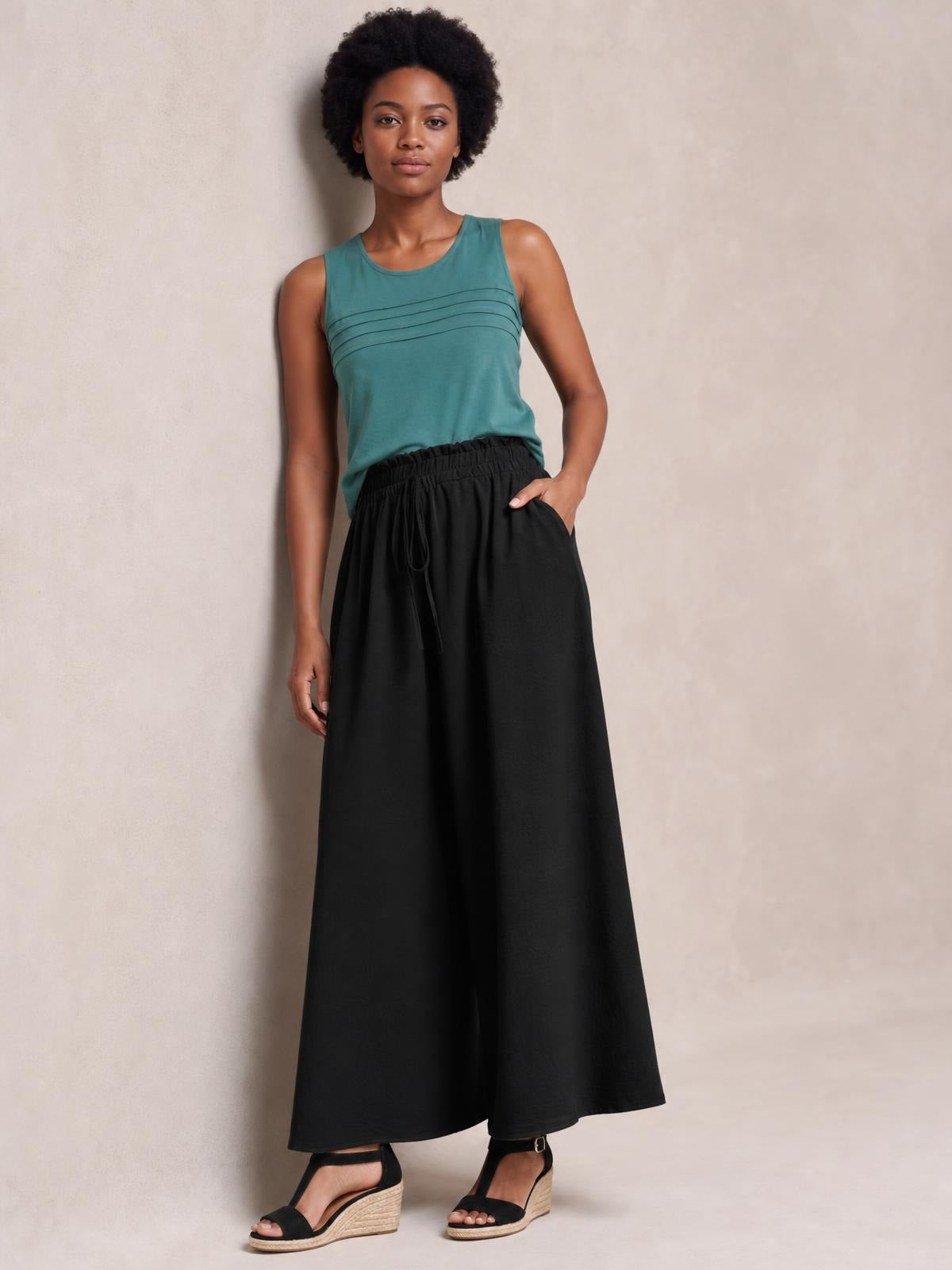 A model poses in black tied high-waist wide-leg pants with an elastic drawstring waistband and flowing opaque silhouette, one hand resting in a pocket.