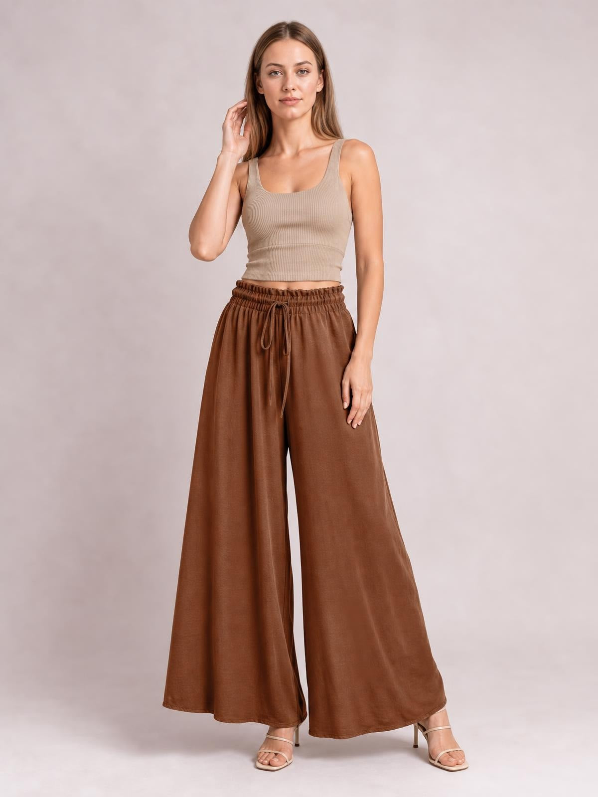 Model wearing brown tied high-waist wide-leg pants with a gathered waistband and flowing silhouette, standing with one hand resting at her side.