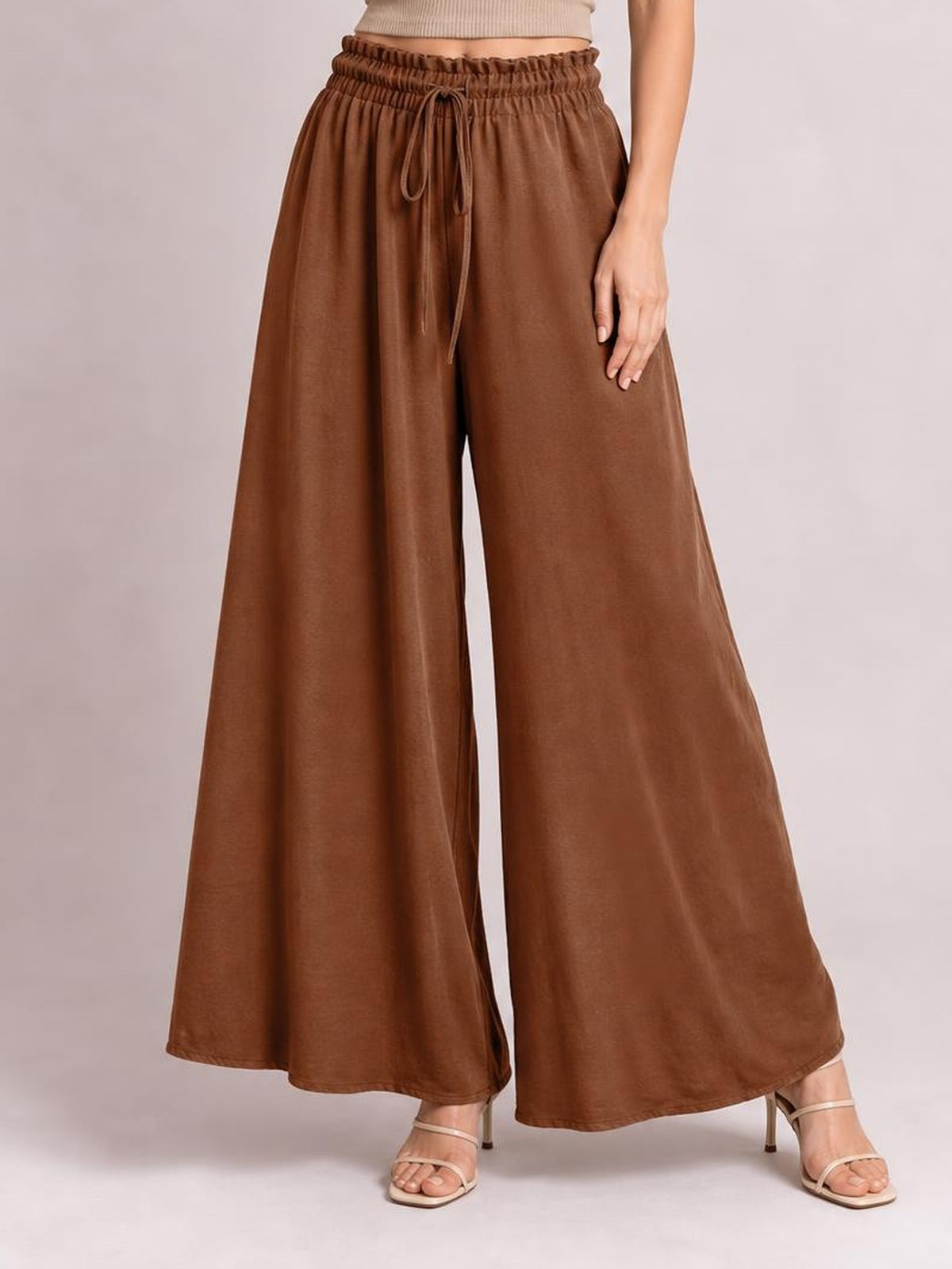 A model wears brown tied high waist wide leg pants with a ruched elastic waistband, flowing opaque fabric, and a relaxed silhouette.