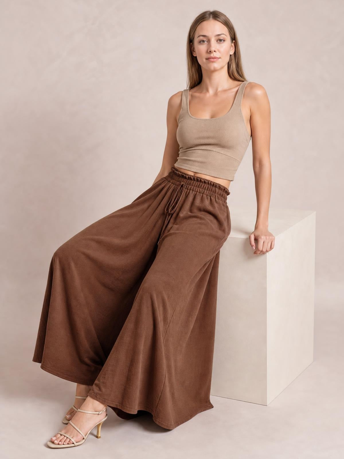 A seated model wears brown tied high-waist wide-leg pants with a relaxed, flowing silhouette, elastic waistband, and visible front drawstring.