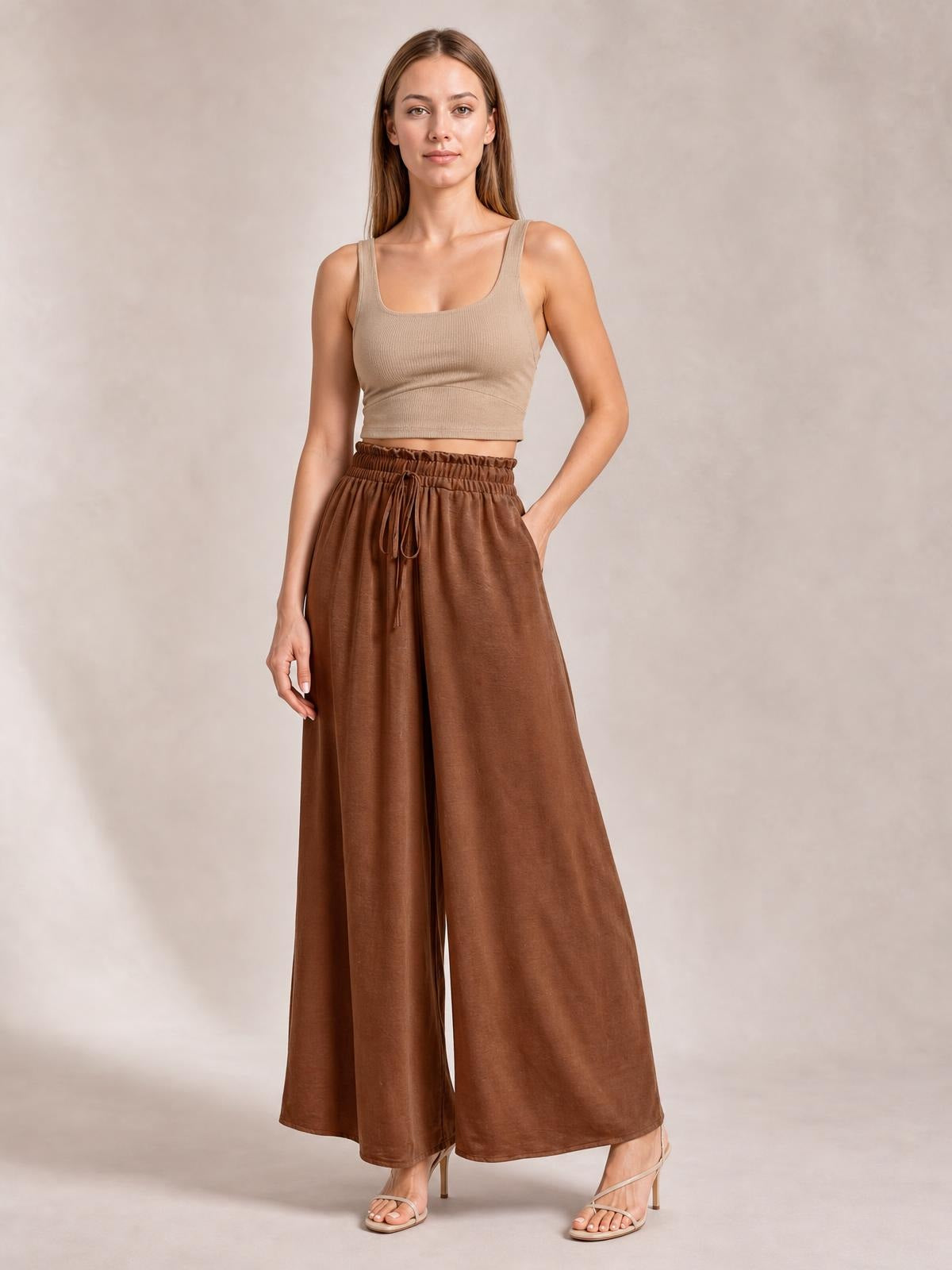 Model poses in brown tied high-waist wide-leg pants with a gathered waistband, drawstring front, flowing silhouette, and cropped beige tank top.