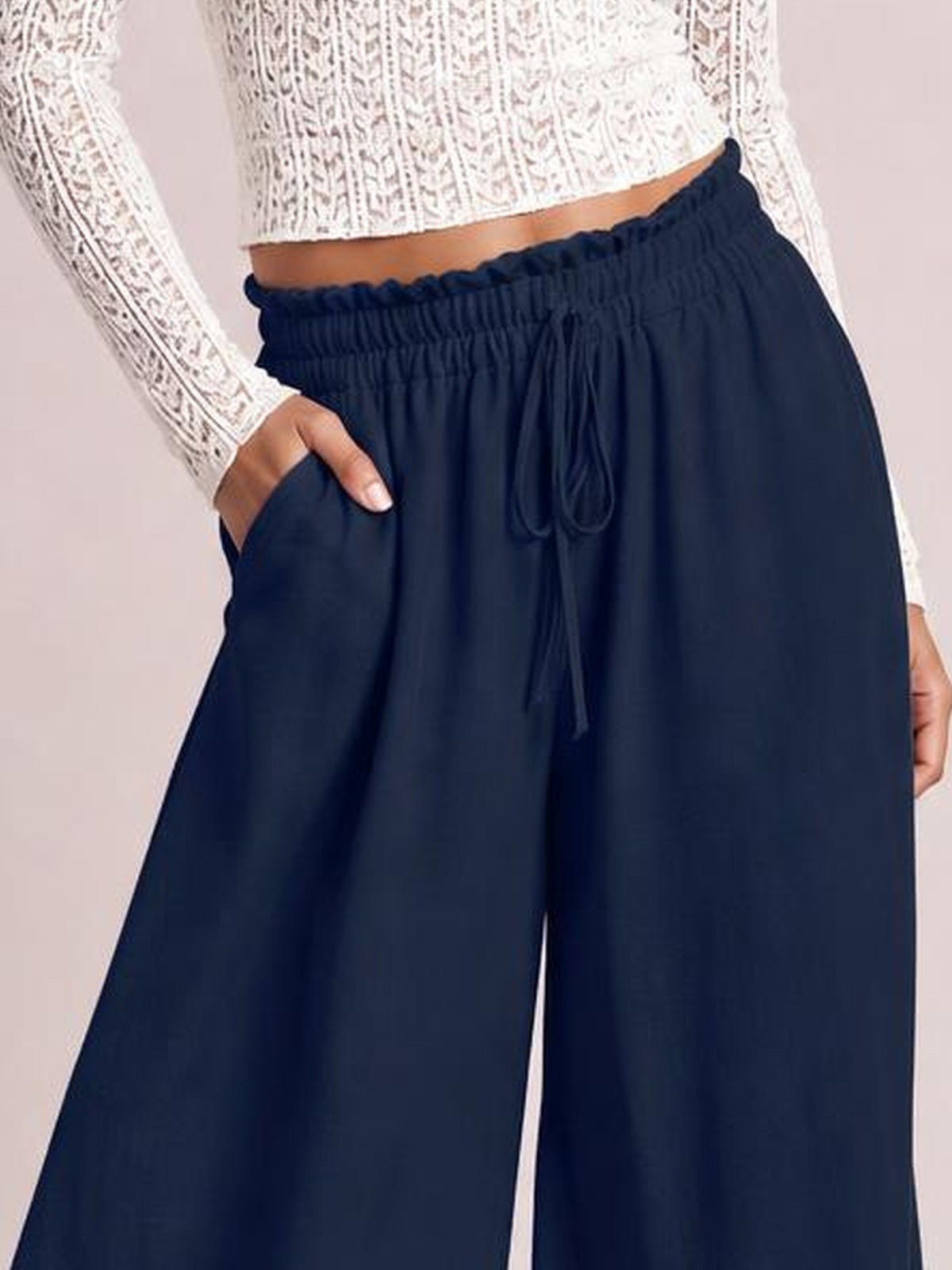 Model wearing navy high-waist wide-leg pants with a gathered elastic waistband, front tie, and side pockets, shown with one hand tucked into a pocket.
