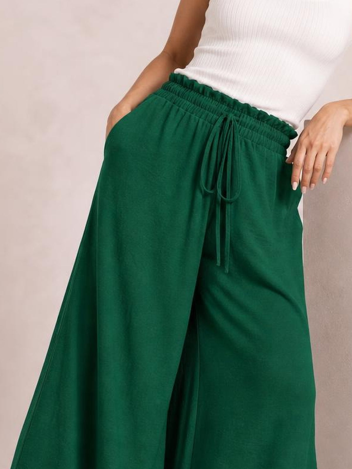 A model poses with one hand in a pocket, showcasing emerald green high-waist wide-leg pants with a gathered waistband and tied drawstring.